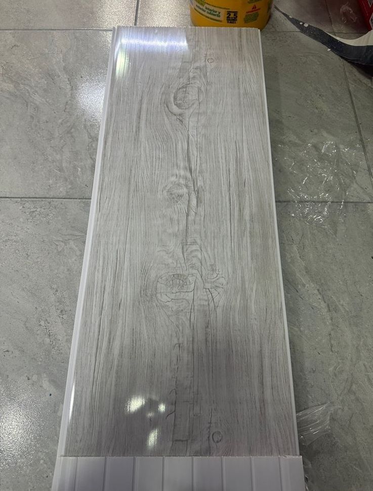 Tablón de suelo de imitación madera gris claro con detalles de veta de madera, sobre una superficie de baldosas grises.