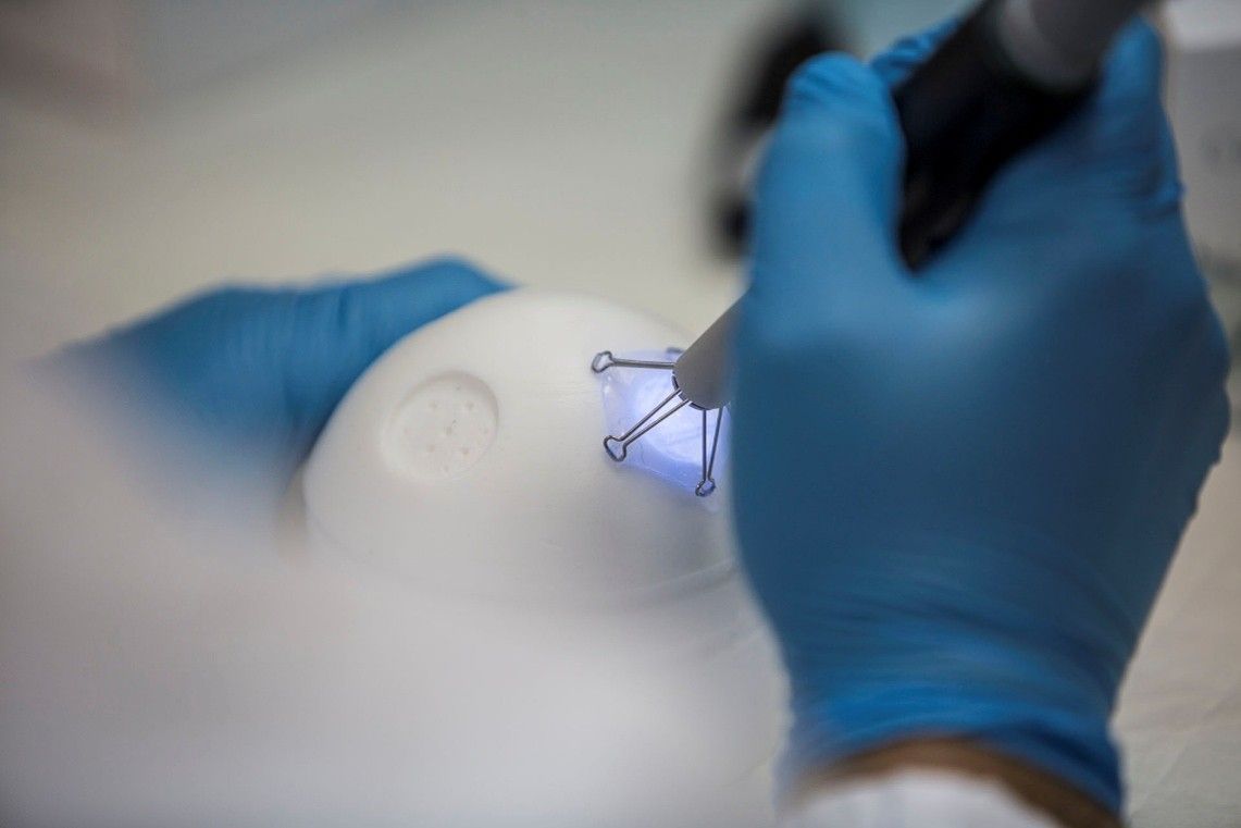 Gloved hands handling a small clear plastic part on a white surface with precision tools