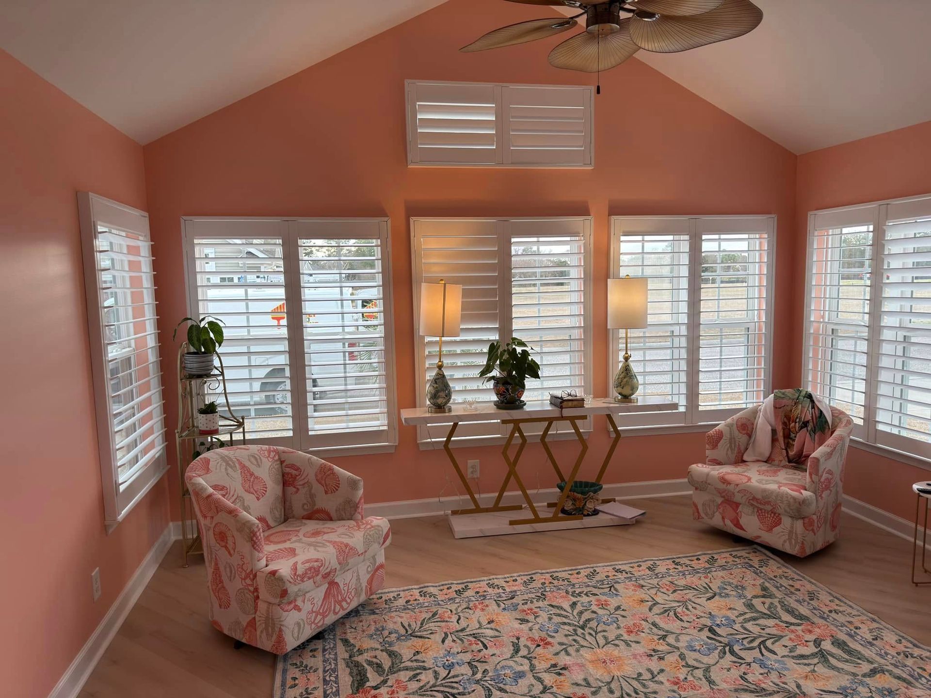 Coral-colored sunroom with floral armchairs, white shutters, and a patterned rug, overlooking a body of water.