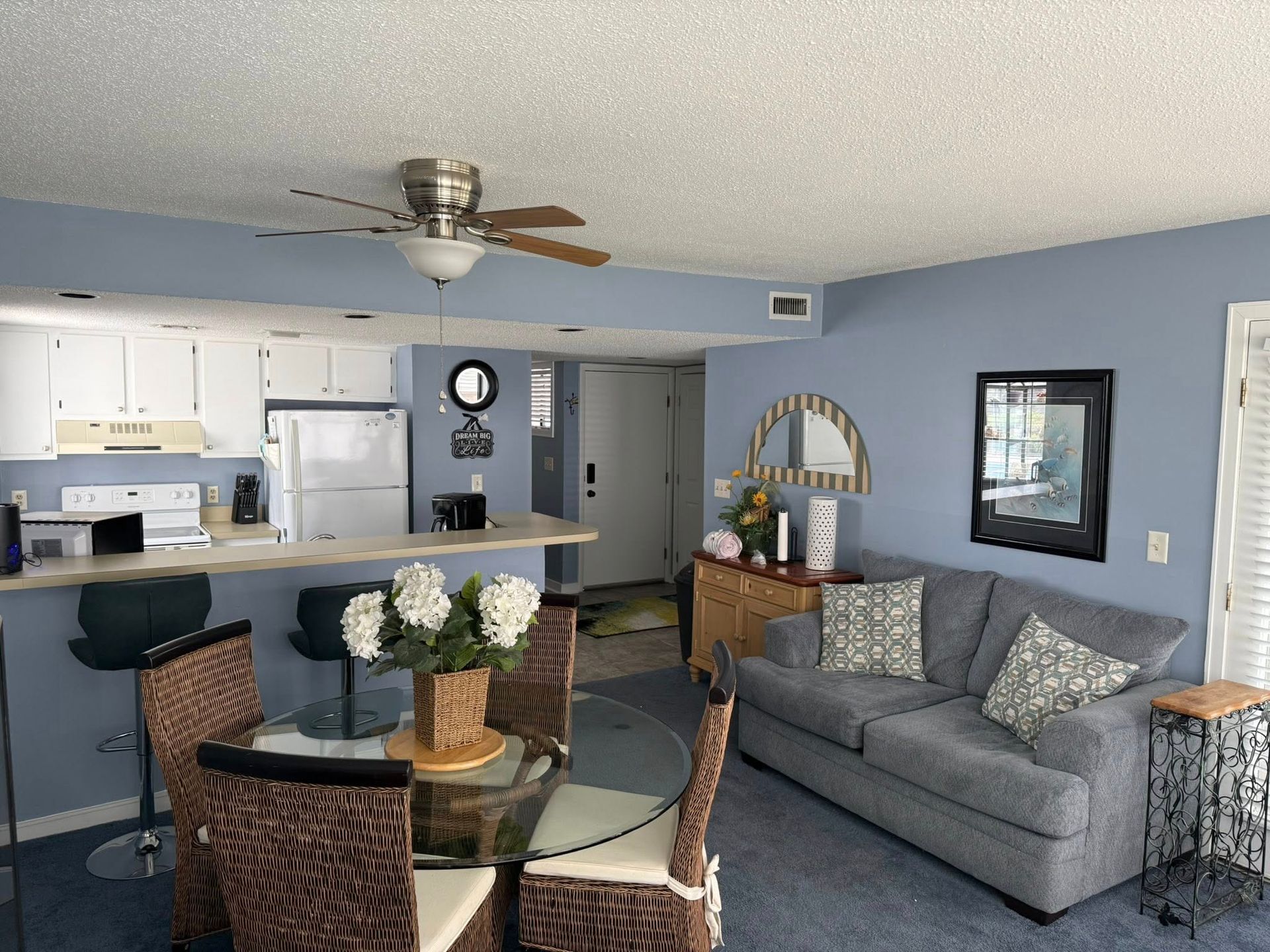 A living room with a dining area, kitchen, and couch. Blue walls and carpet, wicker furniture, and a glass dining table.