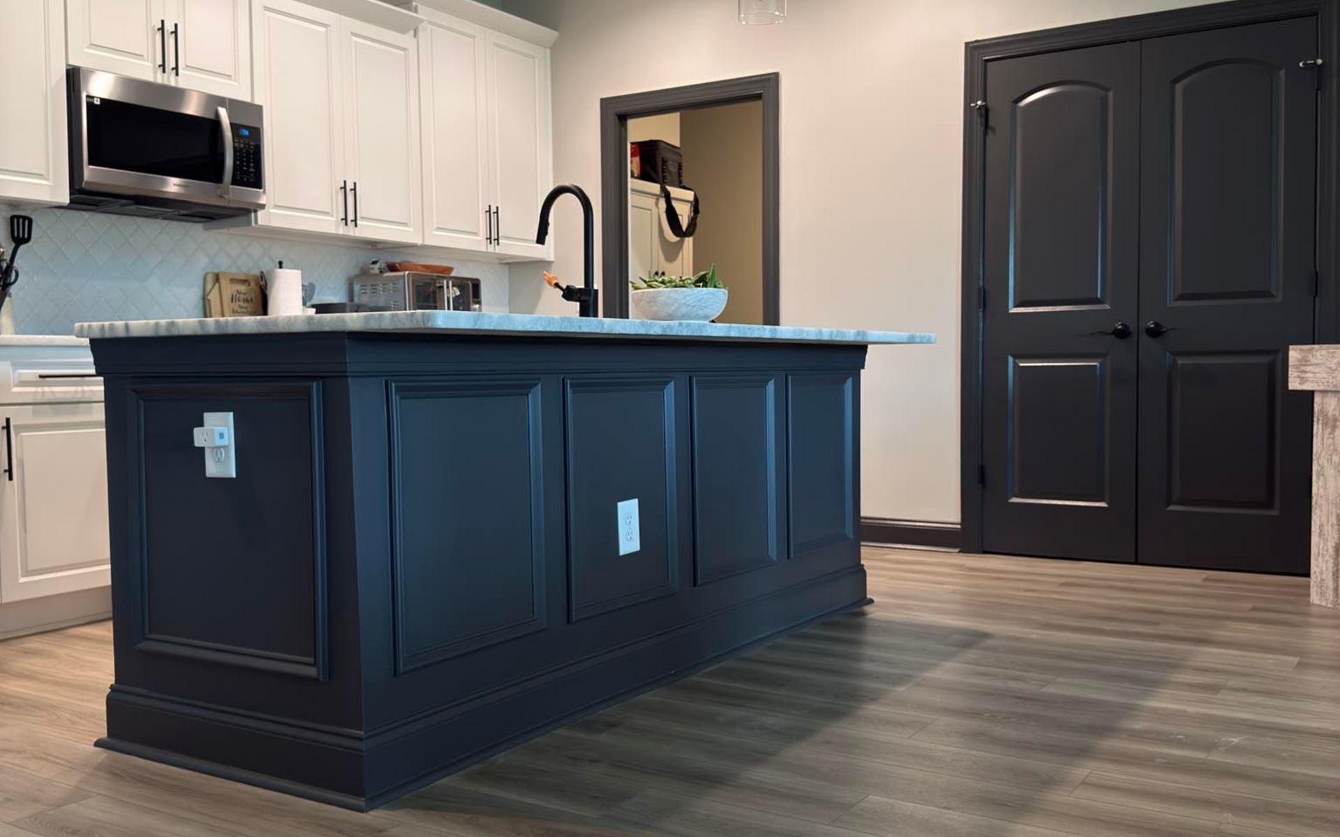 A kitchen island with a dark blue base, marble countertop, and black faucet. White cabinets and a microwave in the background.