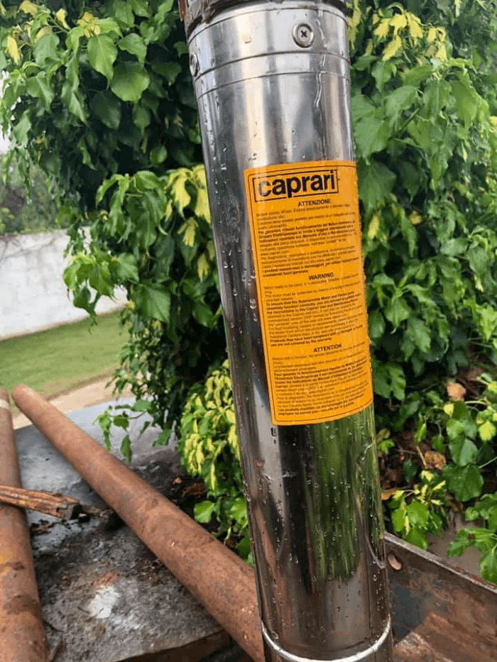 Pompa dell'acqua Caprari in acciaio inox con etichetta gialla del prodotto, all'aperto vicino a vegetazione e tubi arrugginiti.