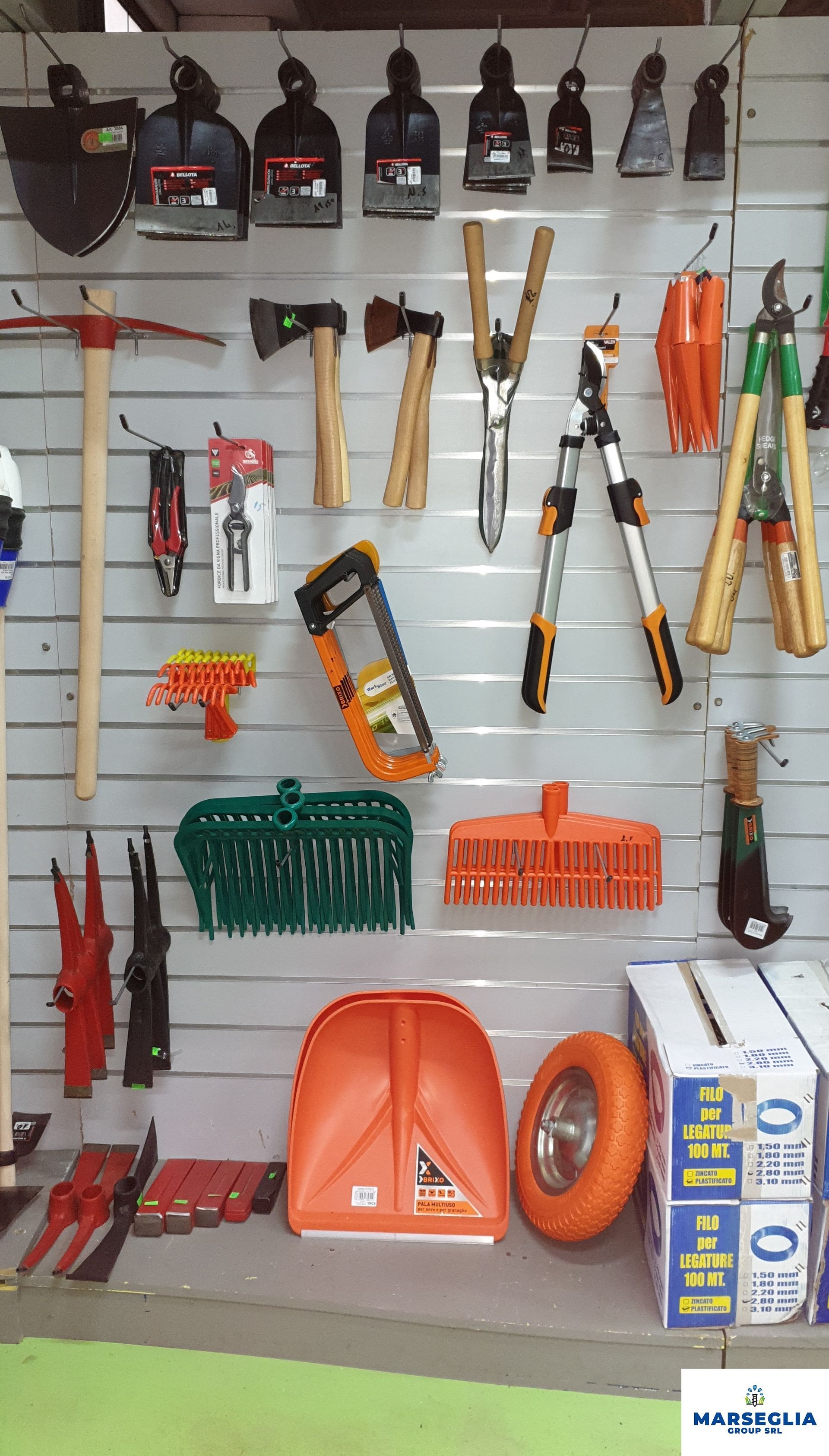 Esposizione di attrezzi in un negozio di ferramenta: zappe, asce, seghe, cesoie, scope e carriole, su una parete di listelli bianchi.