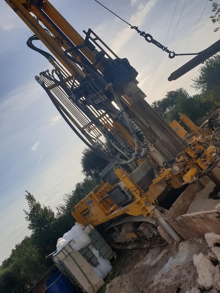 Trapano da cantiere giallo, all'aperto, mentre perfora il terreno. Luce solare, alberi sullo sfondo.