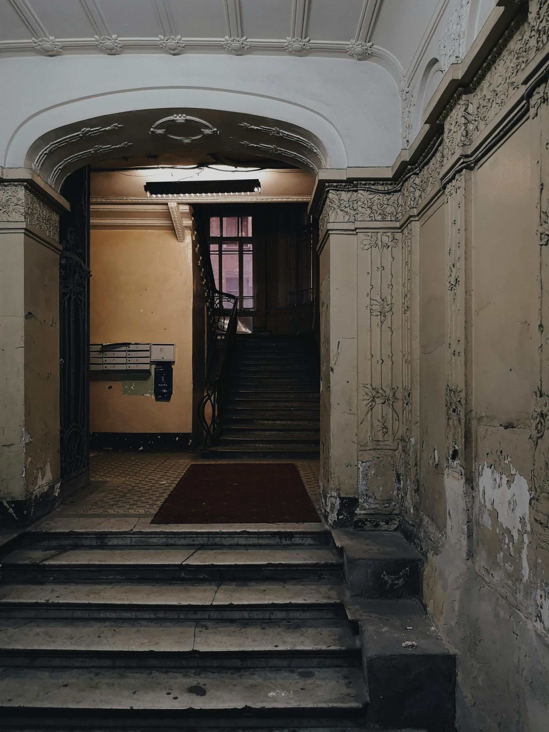 A weathered, ornate entryway with stairs leading towards a dark stairwell and a red carpet.