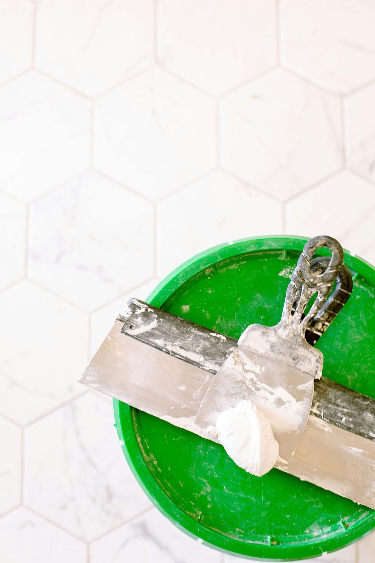 Two putty knives rest on a green bucket, set against a white hexagon tile floor.