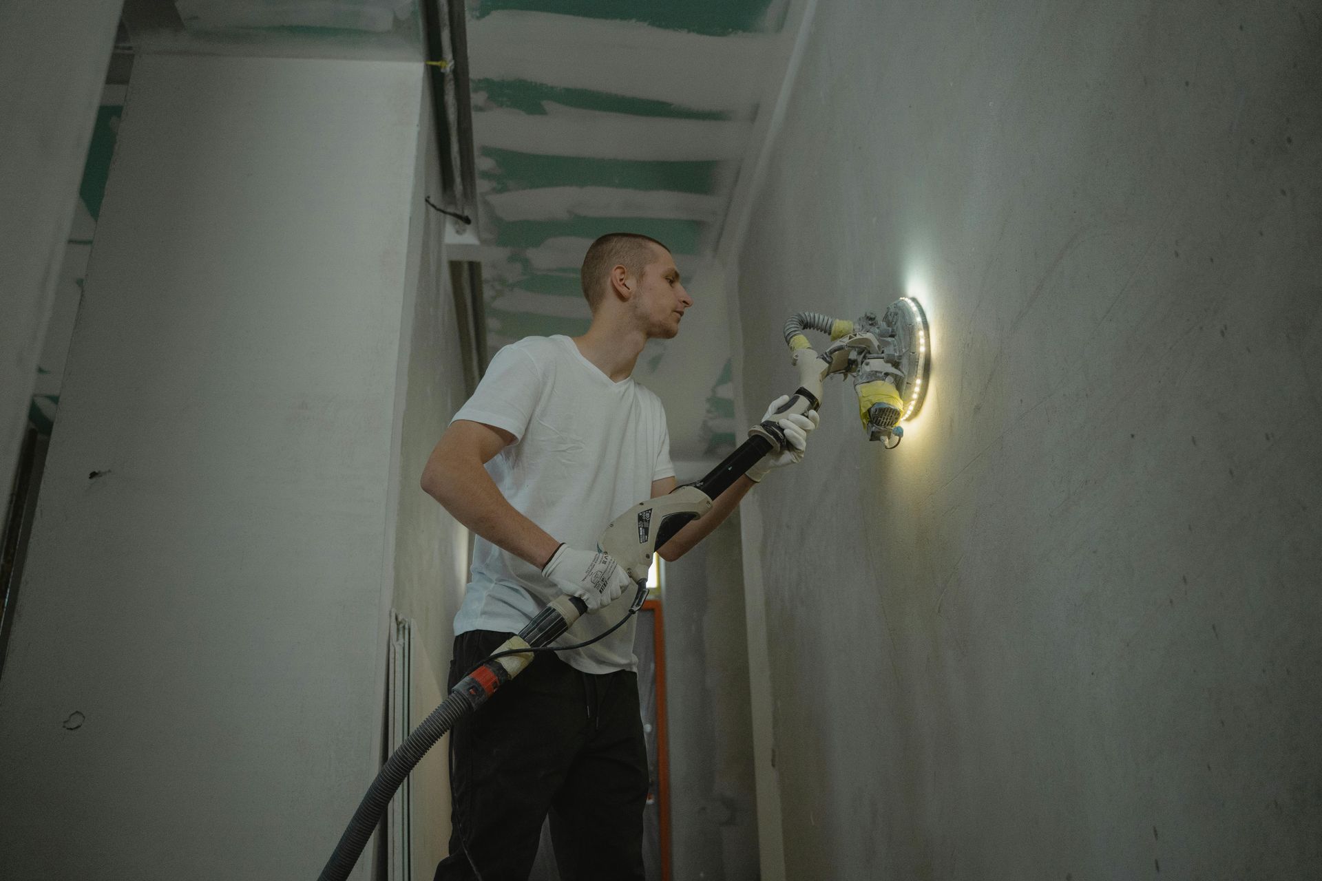 Man sanding a wall with a power sander, wearing gloves and looking focused in a room.