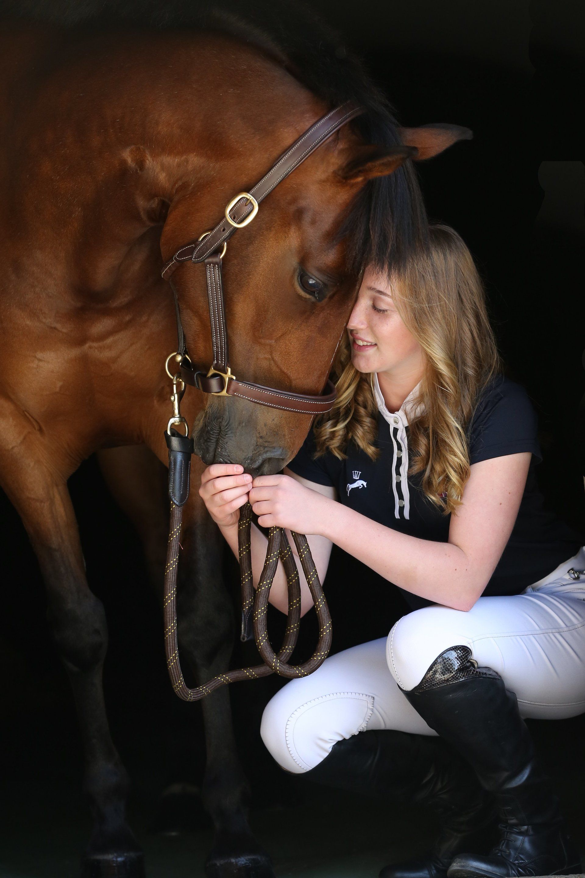 Woman in riding gear crouches, gazing at a brown horse; dark background.