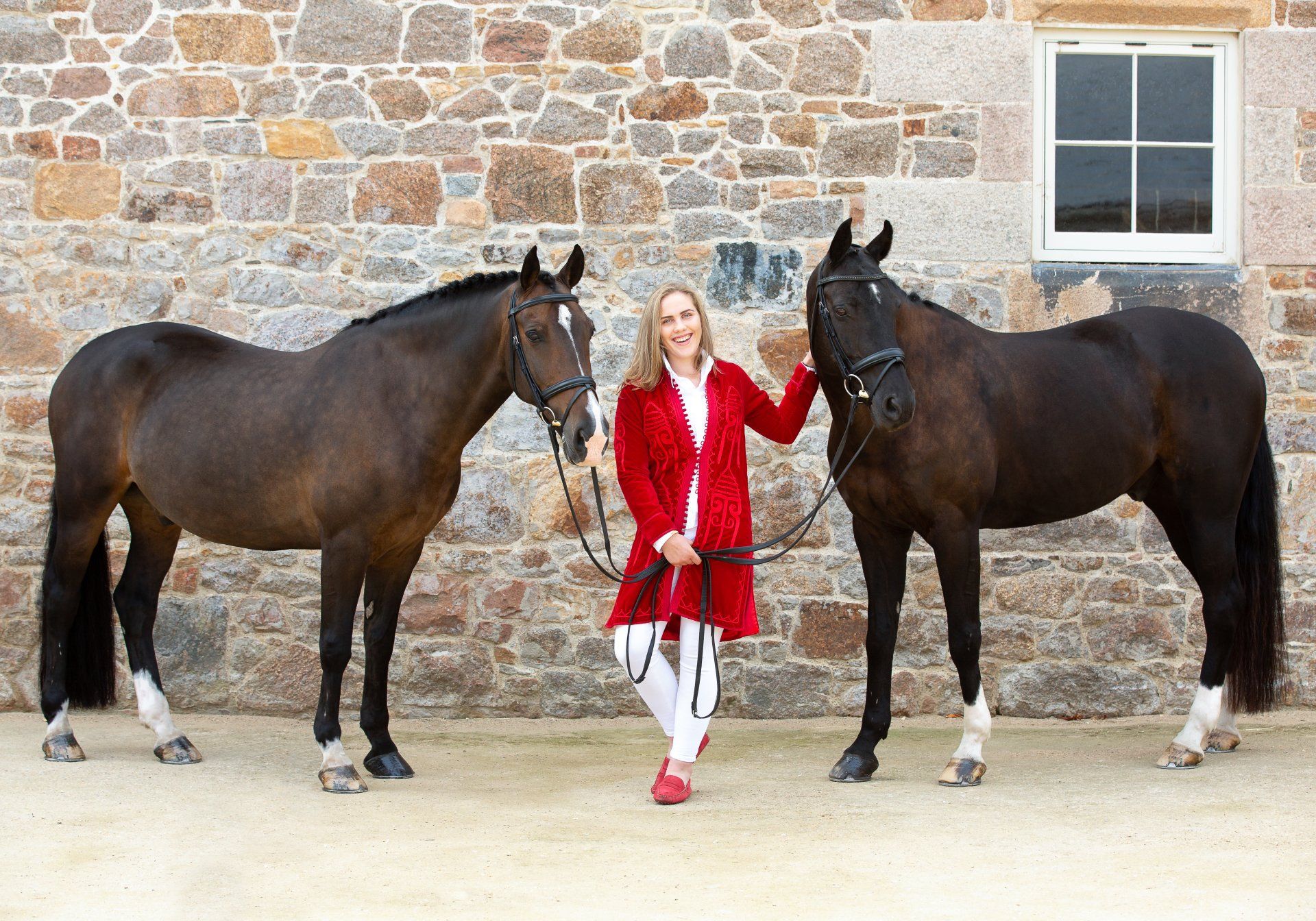 Woman in red coat stands between two dark horses against stone wall.