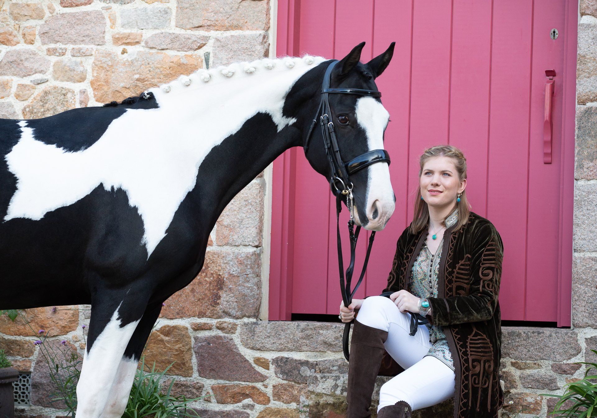 Woman with a black and white horse by a red door; stone wall in background.