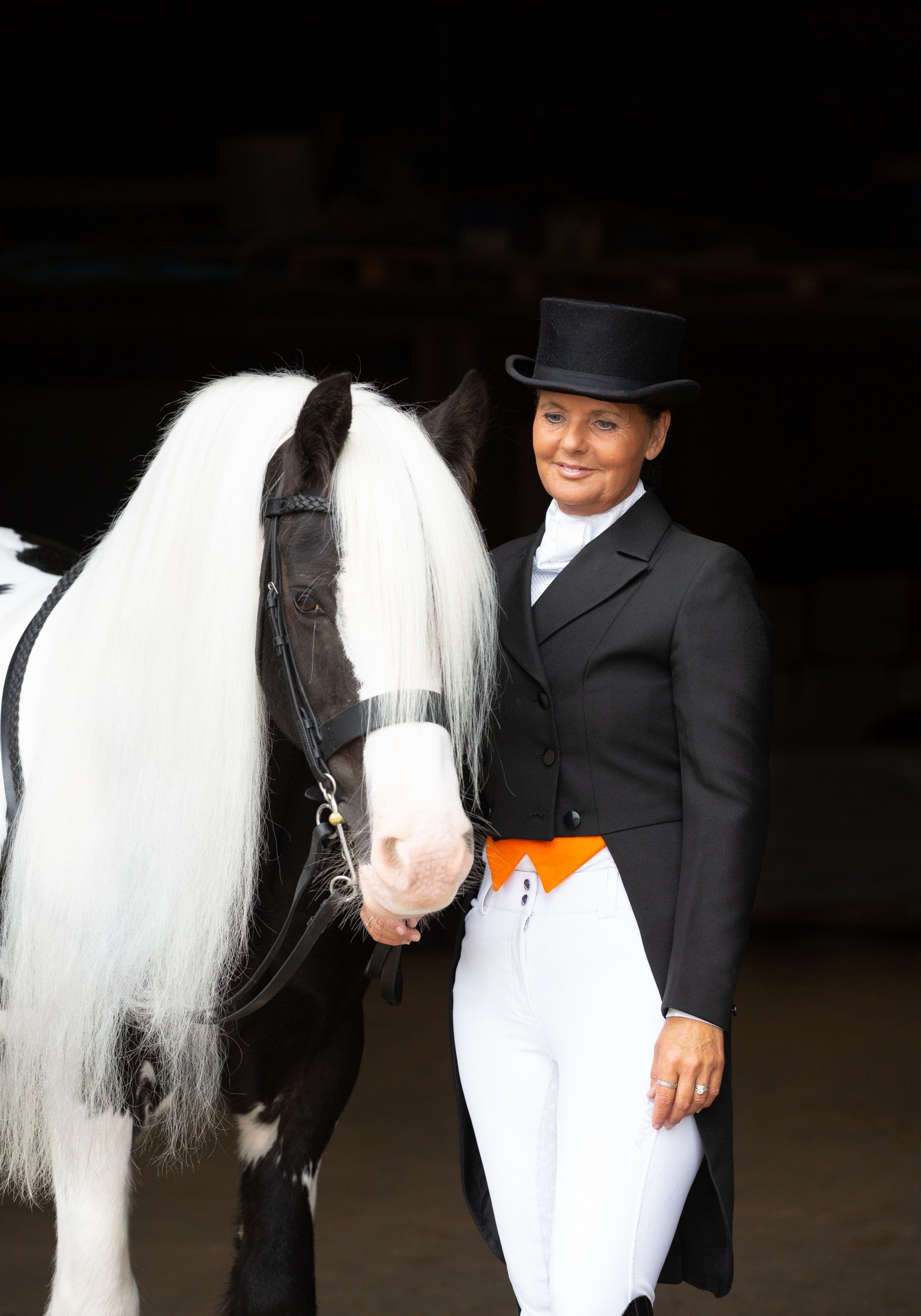 Woman in formal riding attire stands with a black and white horse, posing in a stable.