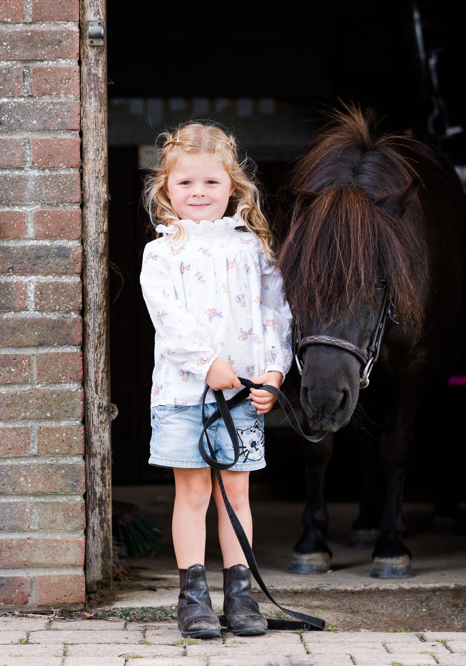 Young child in denim shorts and boots stands with a pony in a stable doorway.