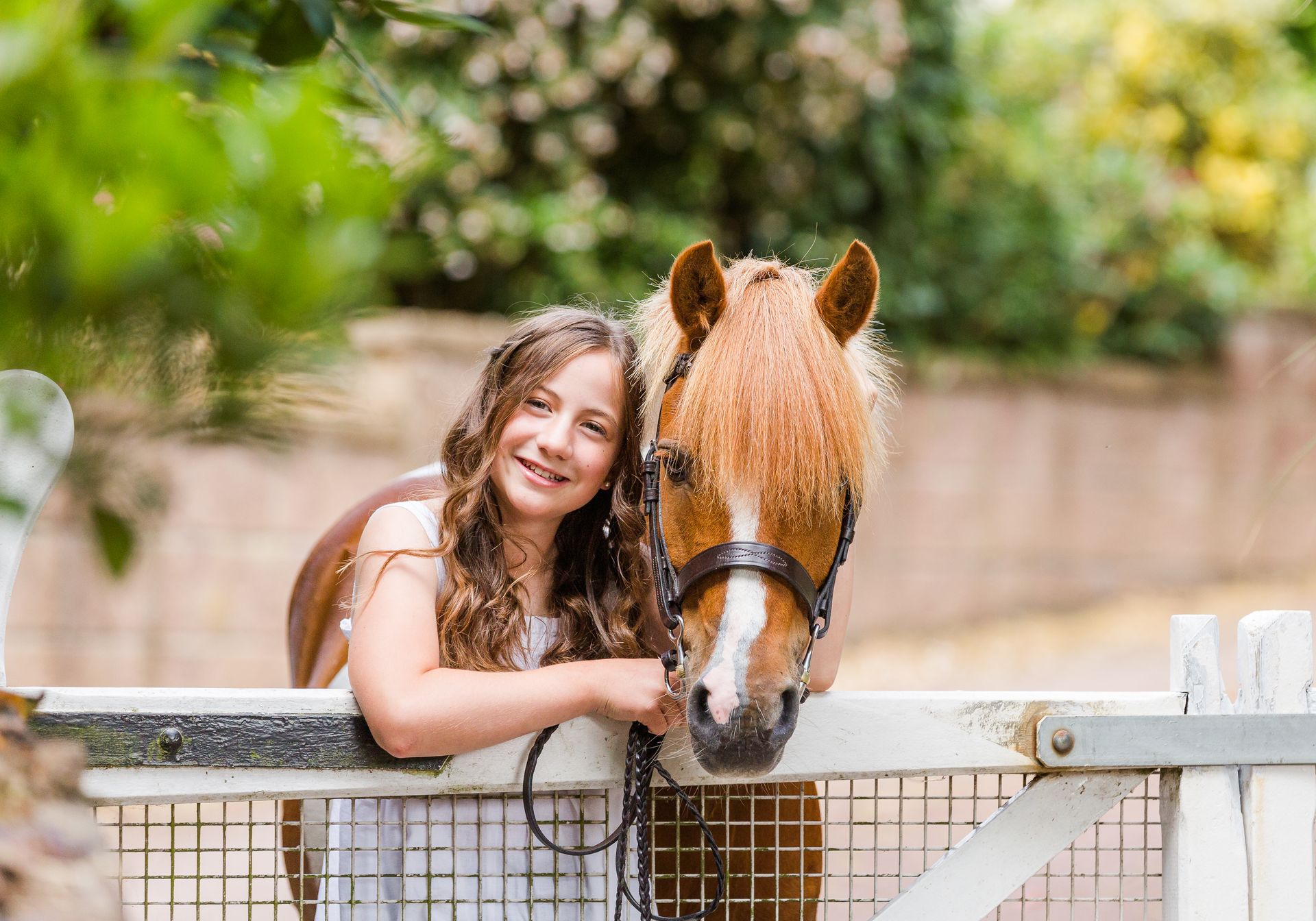 Girl smiles next to a brown pony, leaning on a white fence outdoors.