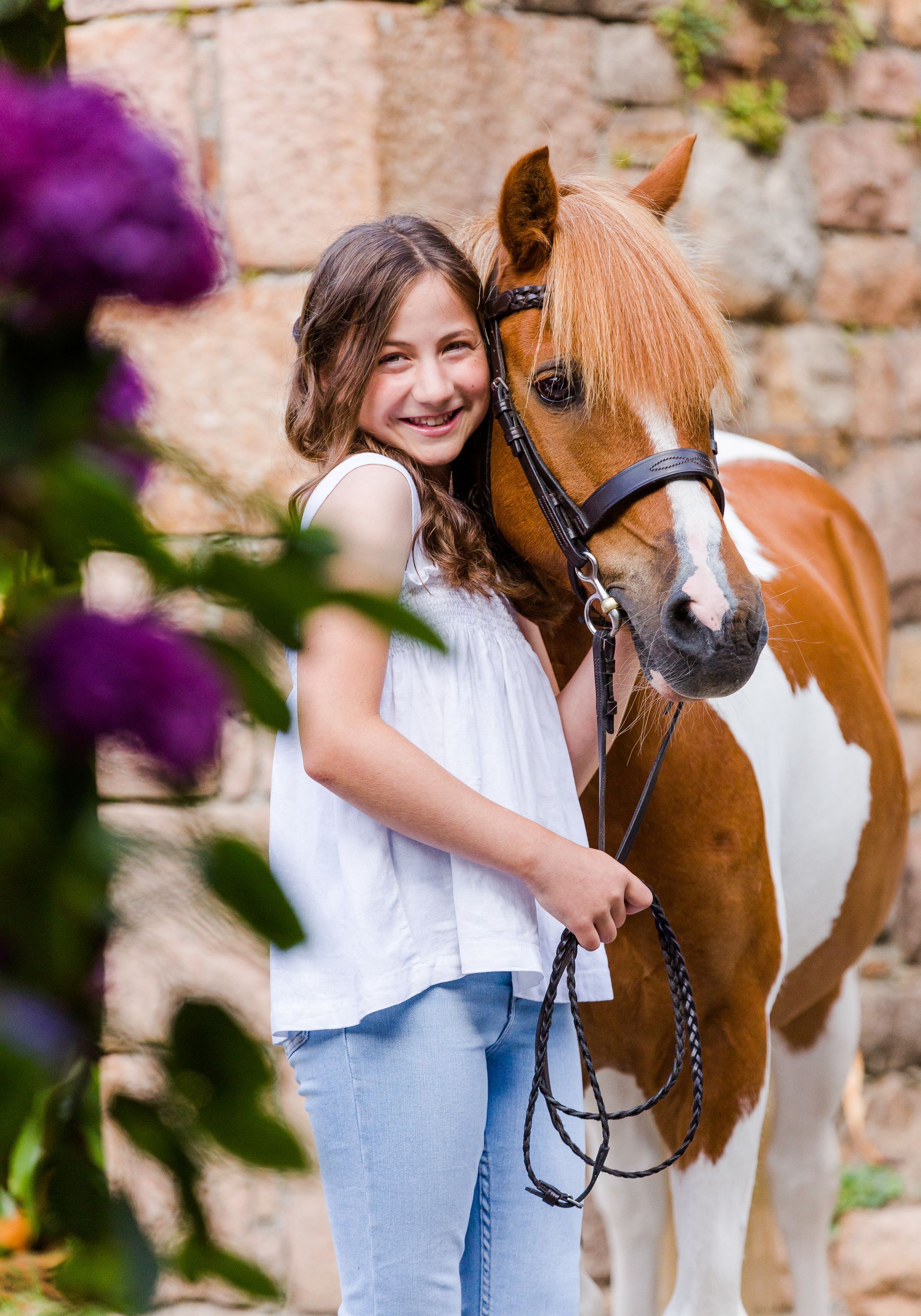 Girl with brown and white pony, smiling, holding reins, standing near purple flowers and stone wall.