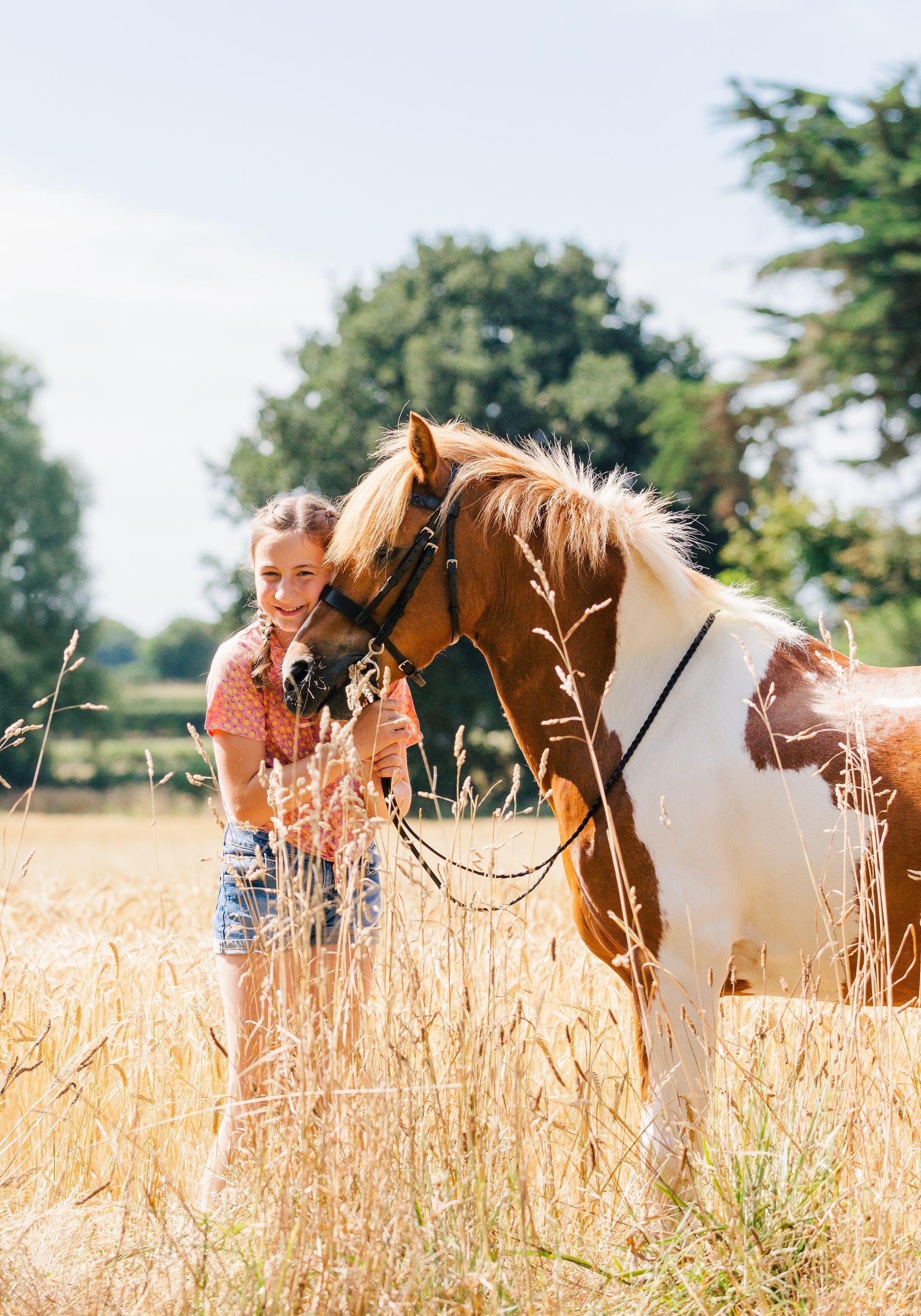Girl petting a brown and white pony in a field of tall grass on a sunny day.