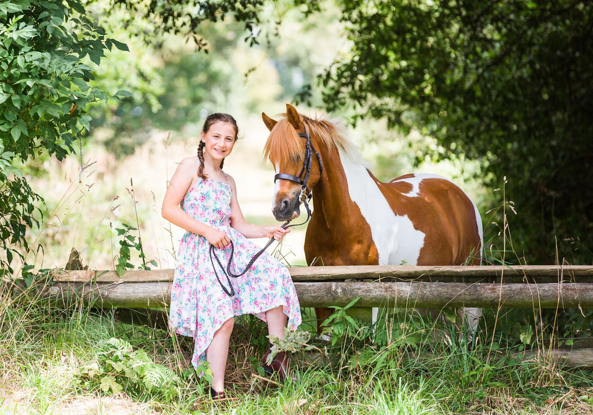Young person in a floral dress sits by a brown and white pony, holding its reins near a wooden fence outdoors.