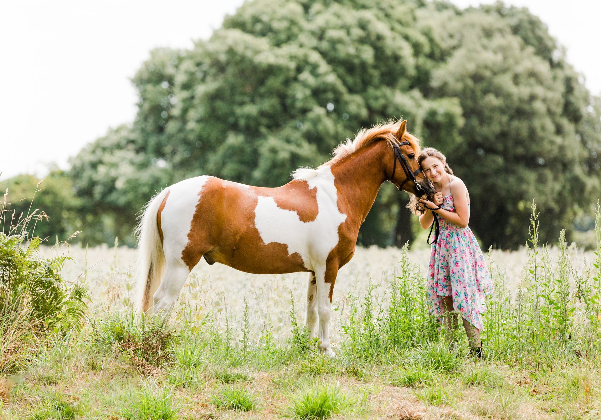 A brown and white pony stands with a woman in a floral dress in a grassy field.