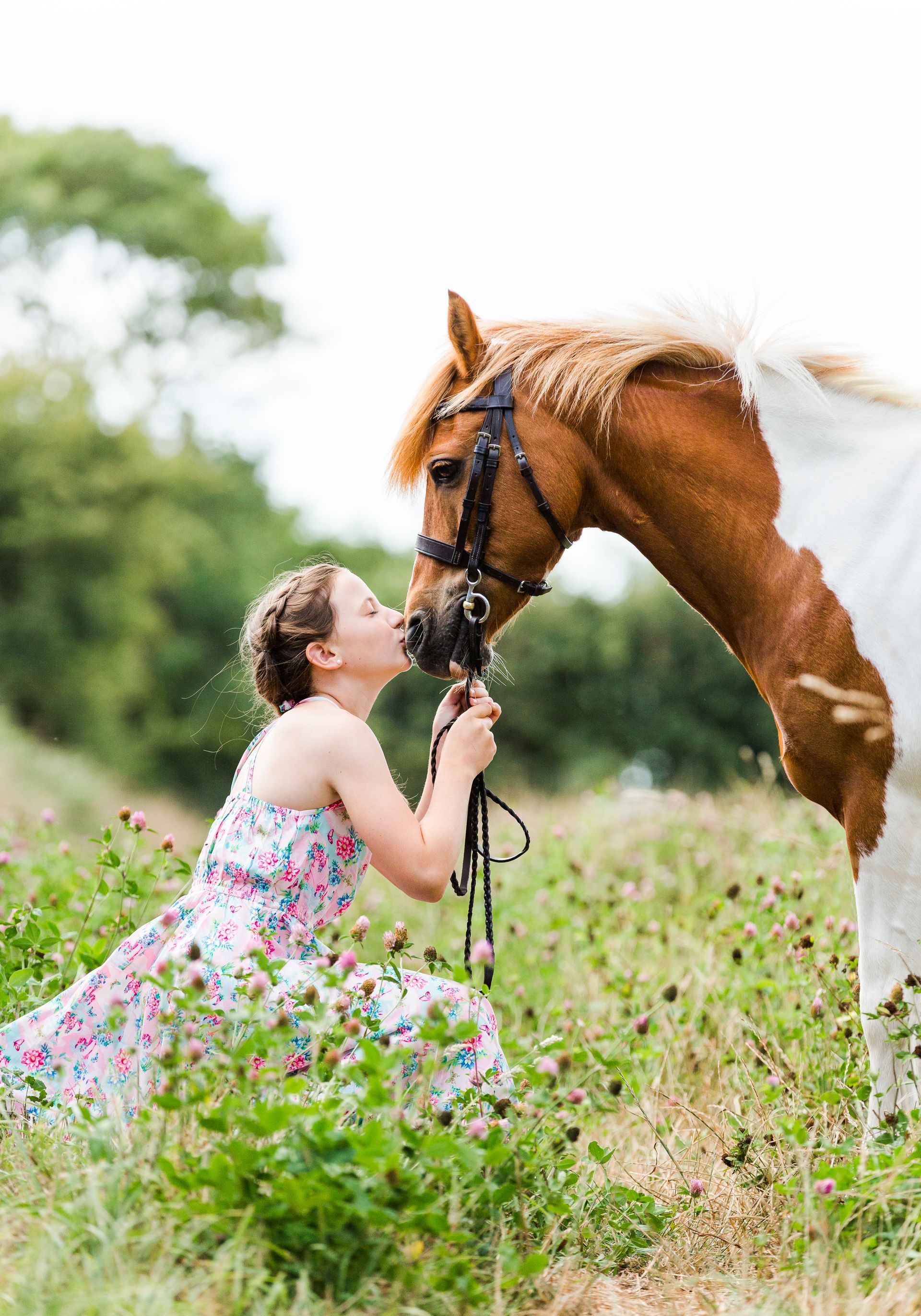 Girl in floral dress kisses horse in a grassy field.