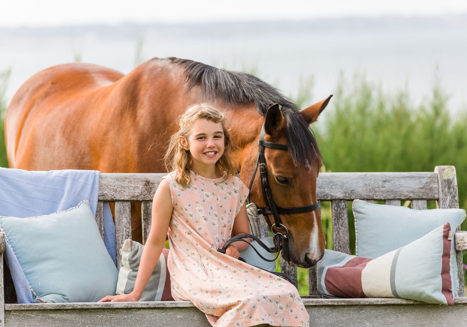 Girl in a pink dress smiles, sitting on bench with a brown horse in a green field with blue pillows.