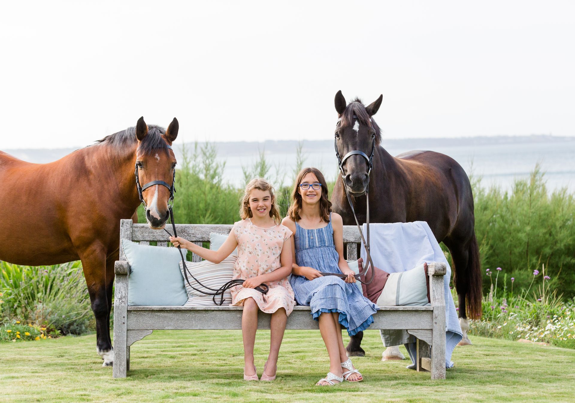 Two girls sit on a bench with two horses on either side; outside, waterfront view.
