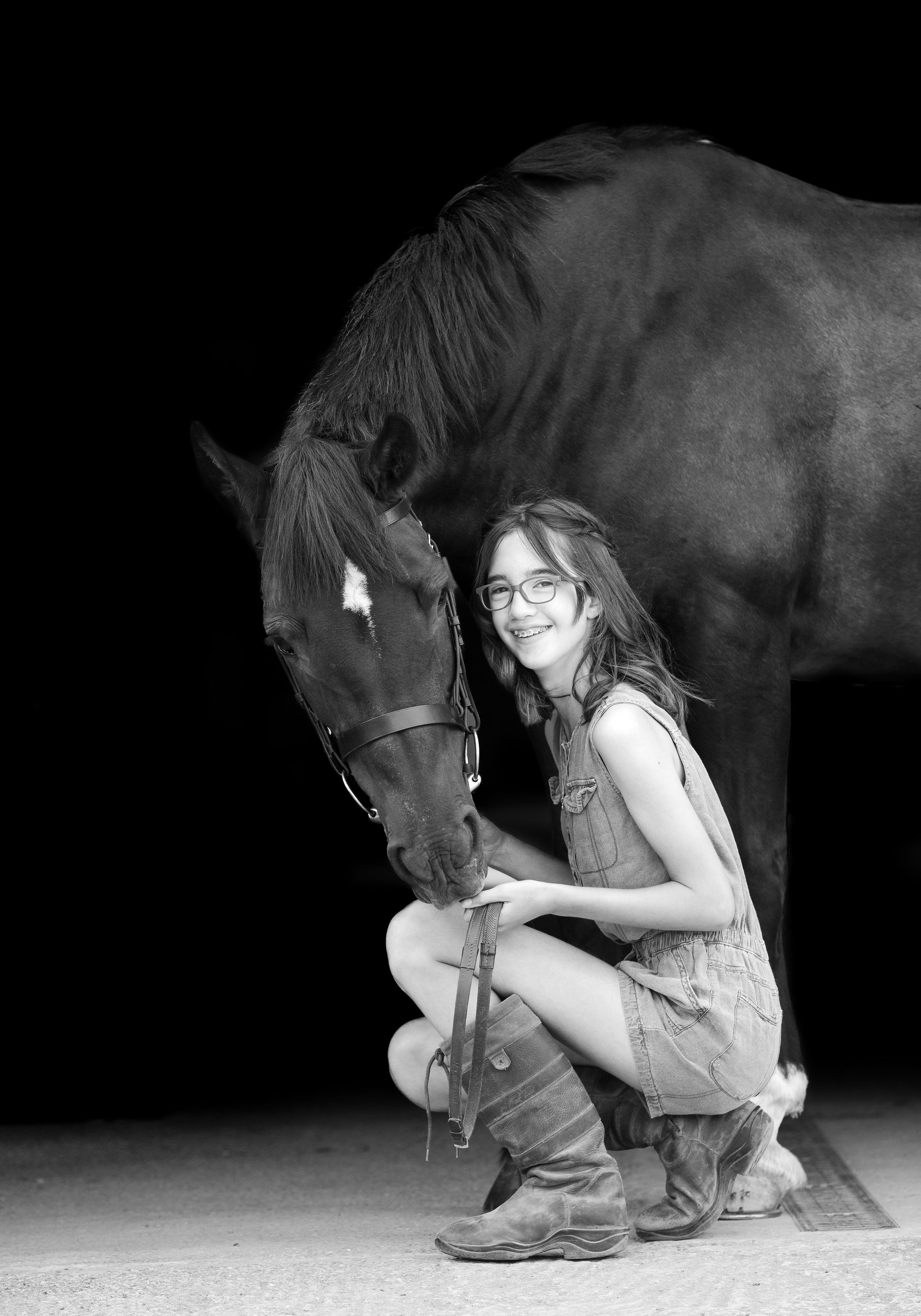 Girl with glasses crouches beside a dark horse in a stable, petting the horse. Black and white.
