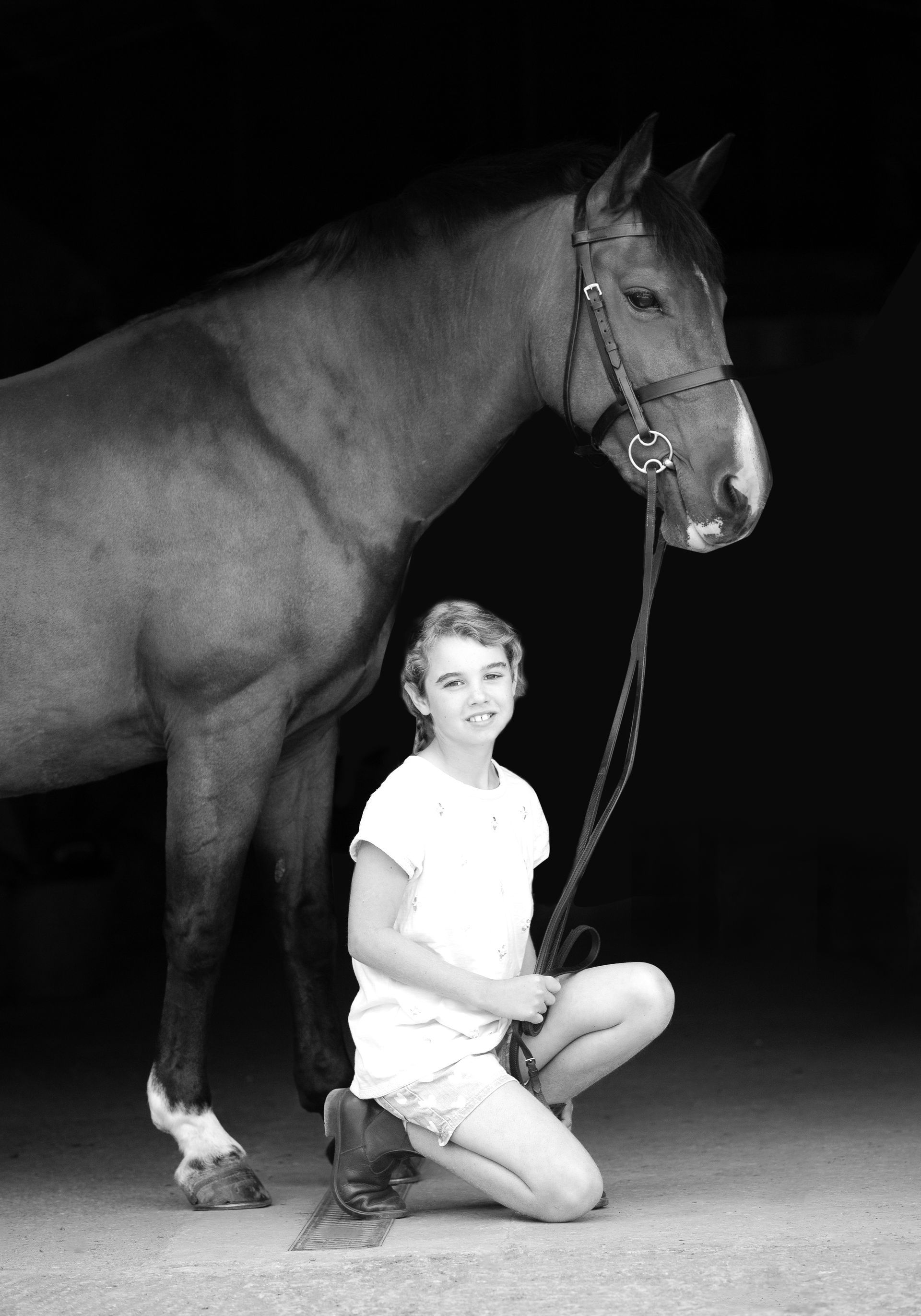 A girl kneels beside a horse. The horse wears a bridle. Both are in front of a dark backdrop.