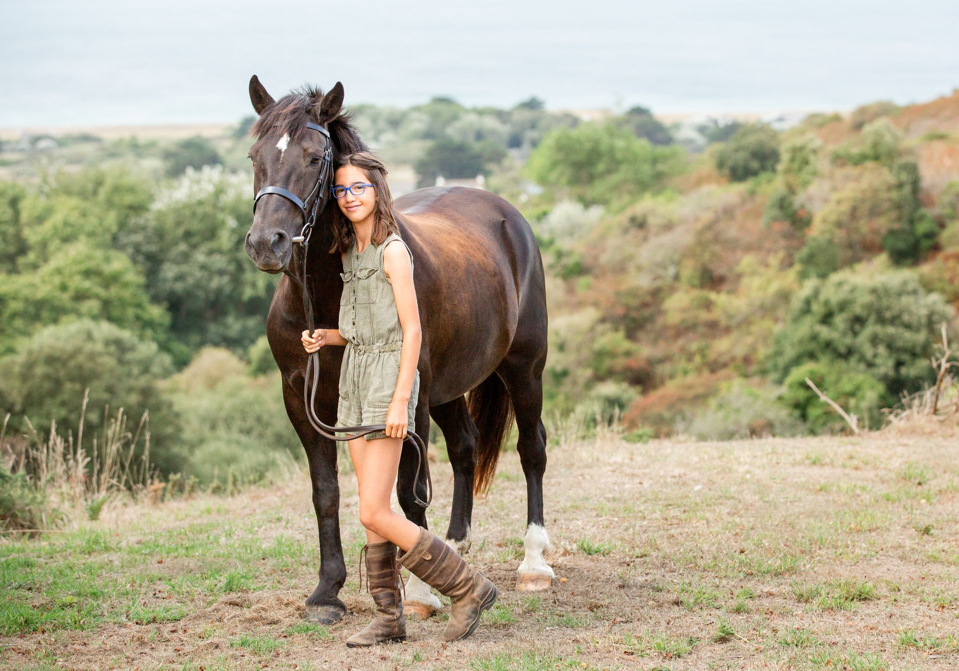 Girl with glasses and a brown horse on a grassy hill; trees and water in the background.