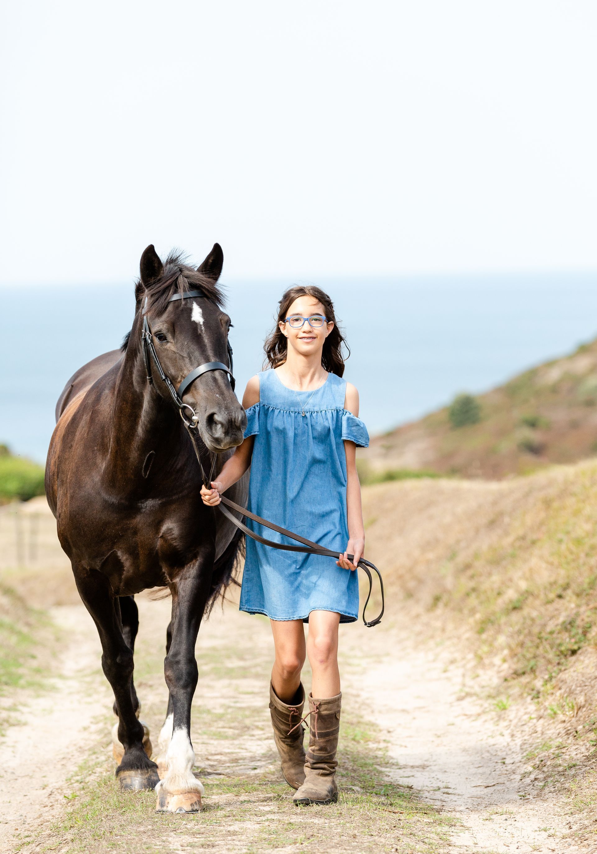 Girl in blue dress and boots walking alongside a dark horse on a dirt path, ocean in the background.