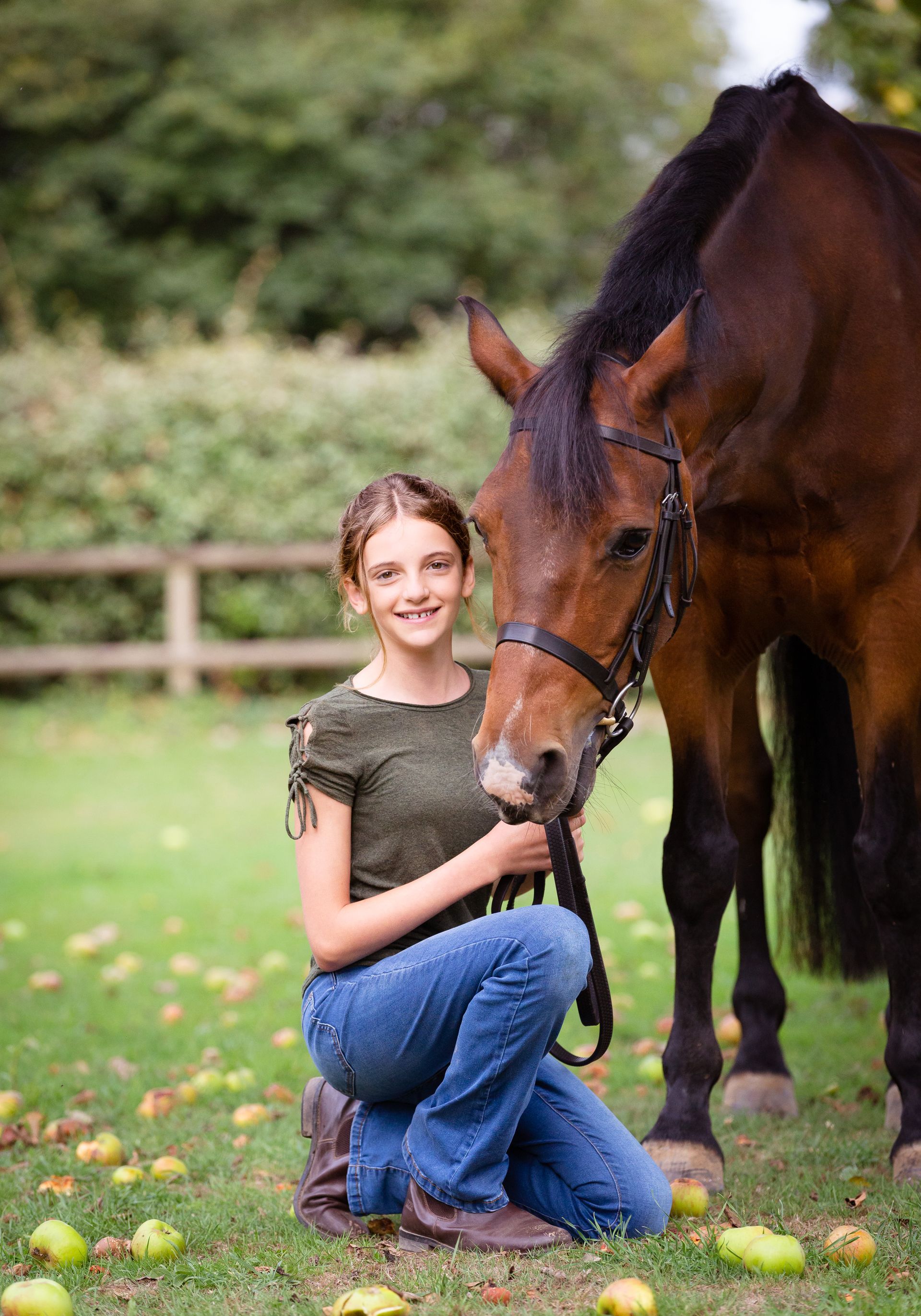 Girl in jeans kneels next to a brown horse in a grassy field, smiling and holding the reins.