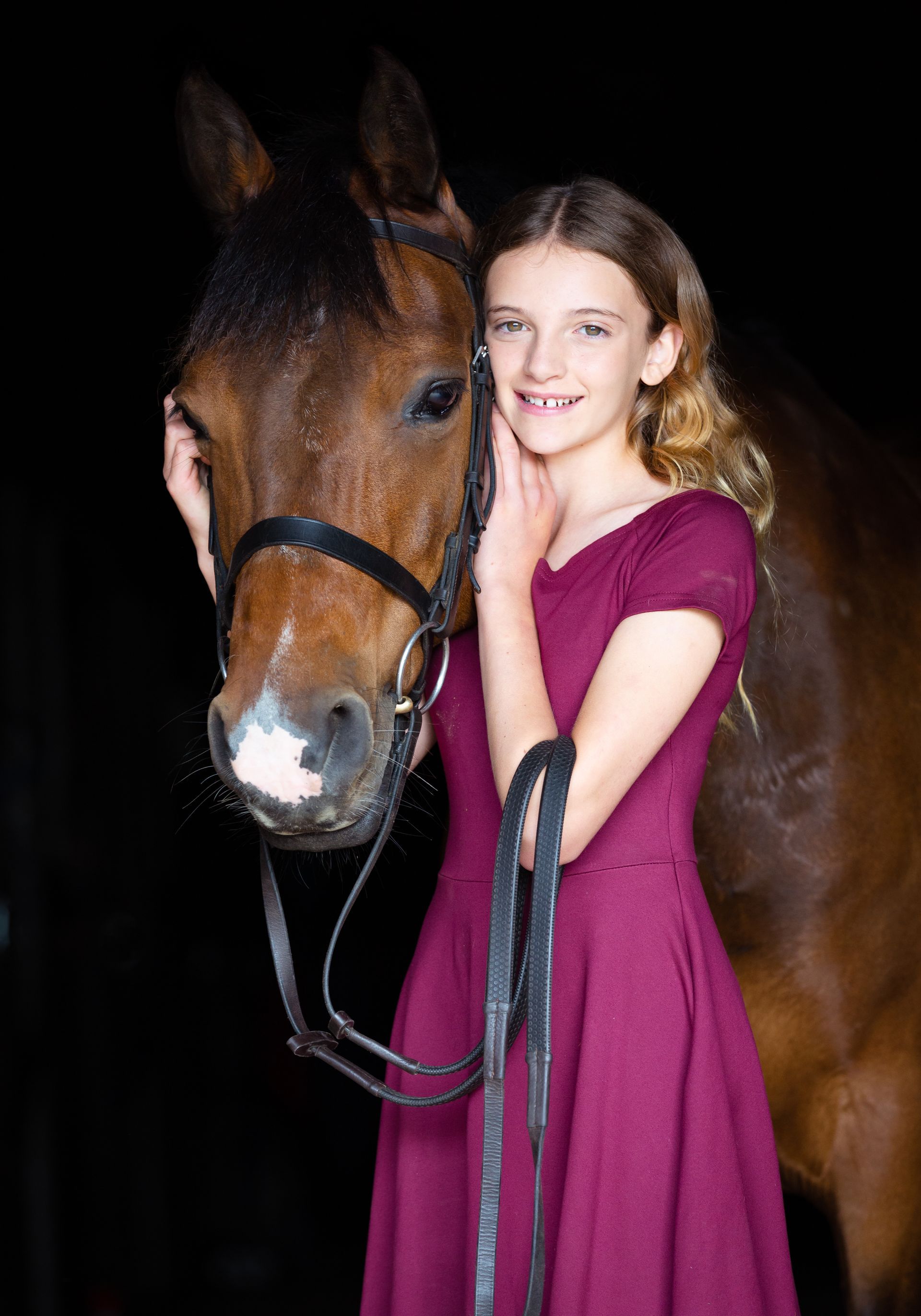 Girl in maroon dress with horse, smiles while gently touching horse's head; dark background.