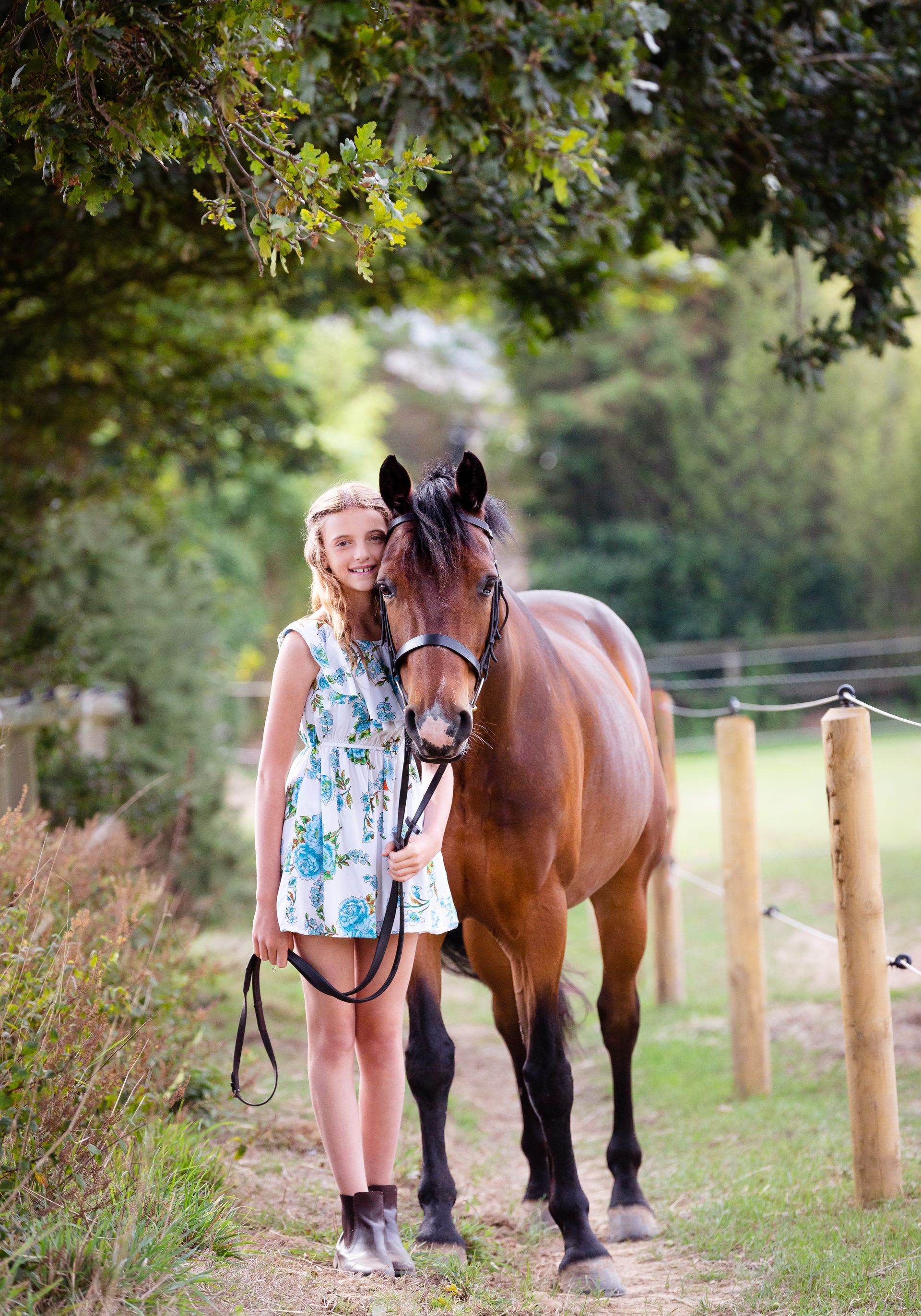 Woman in floral dress standing beside a brown horse, both looking at camera, outdoors.