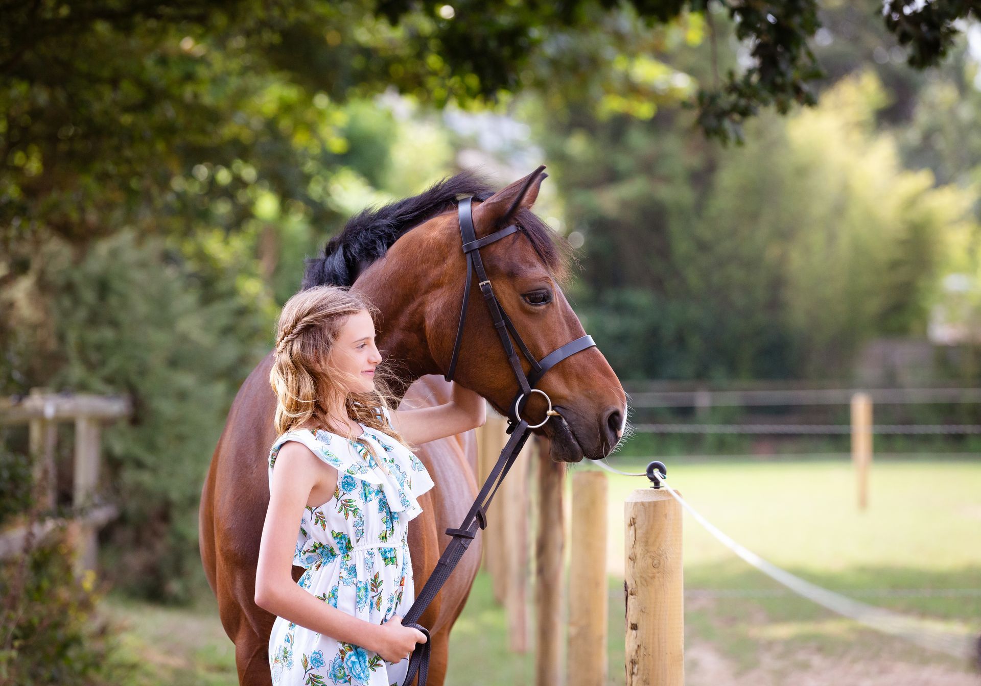 Woman in floral dress with brown horse, standing near a fence in a sunlit outdoor setting.