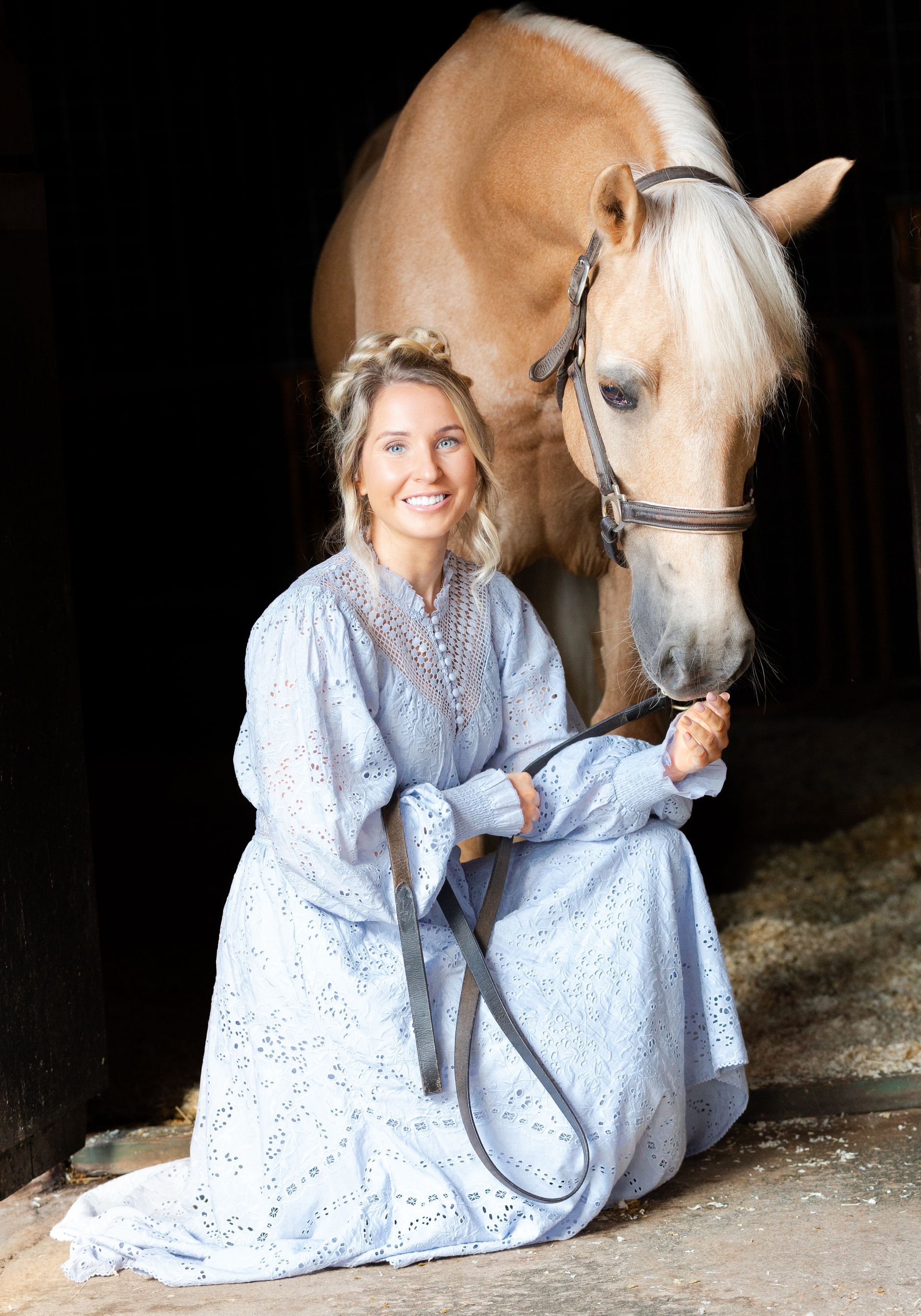 Woman in blue dress kneels beside a tan horse in a stable, smiling and holding the reins.