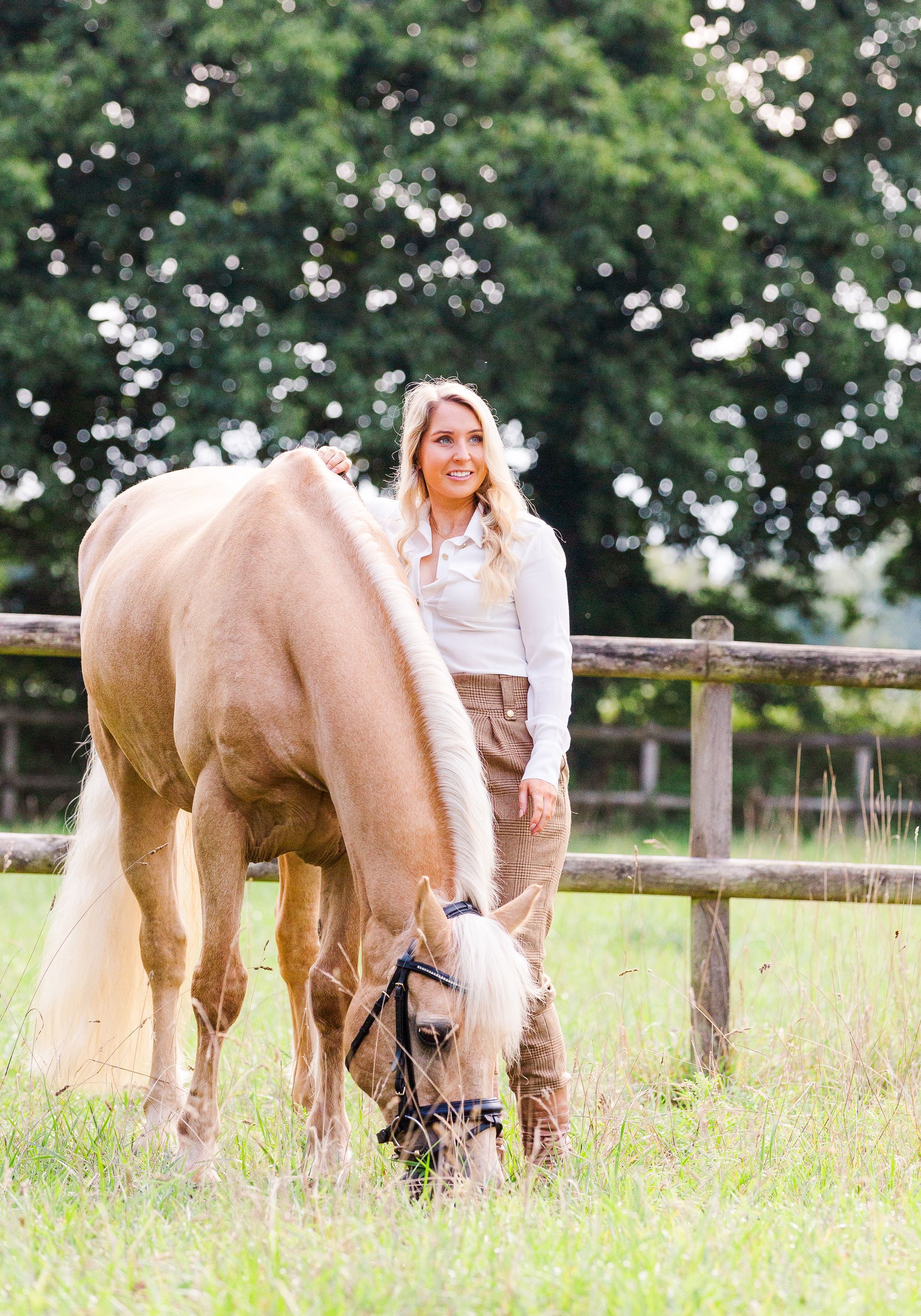 Woman in white shirt and patterned pants standing next to a palomino horse grazing in a grassy field.