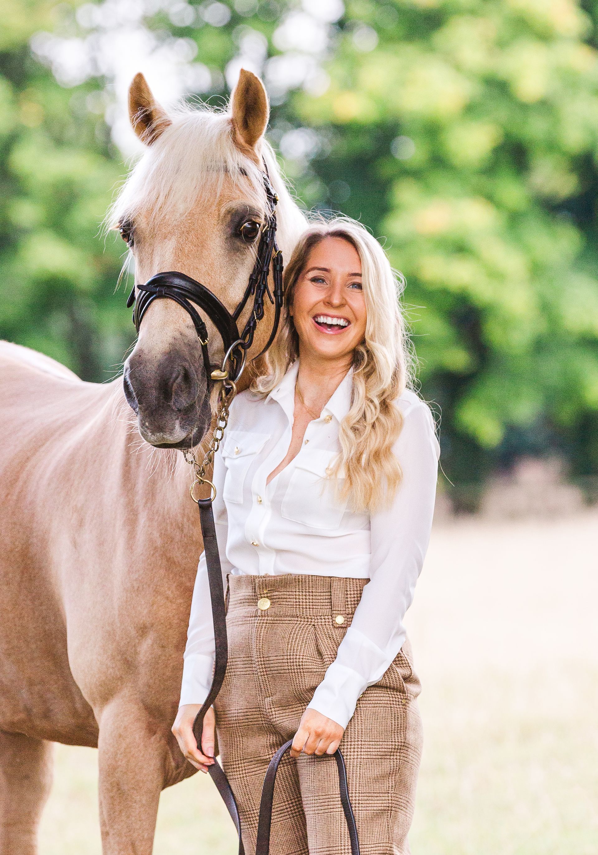 Woman with long blonde hair smiles next to a light brown horse. She wears a white shirt, brown pants, in a green field.