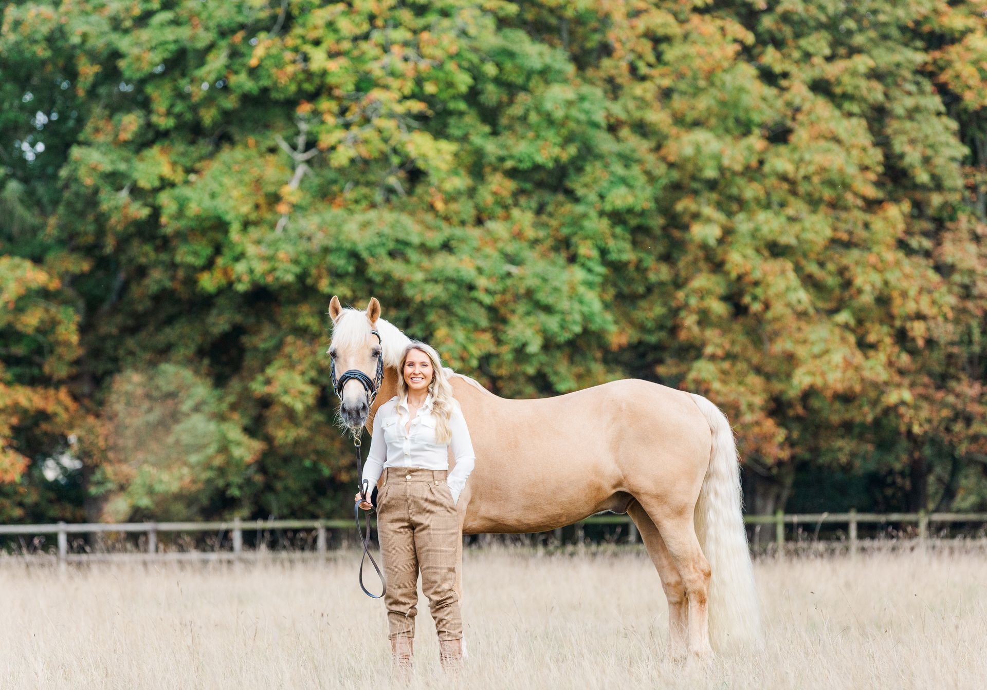 Woman stands next to a palomino horse in a field; trees in the background.