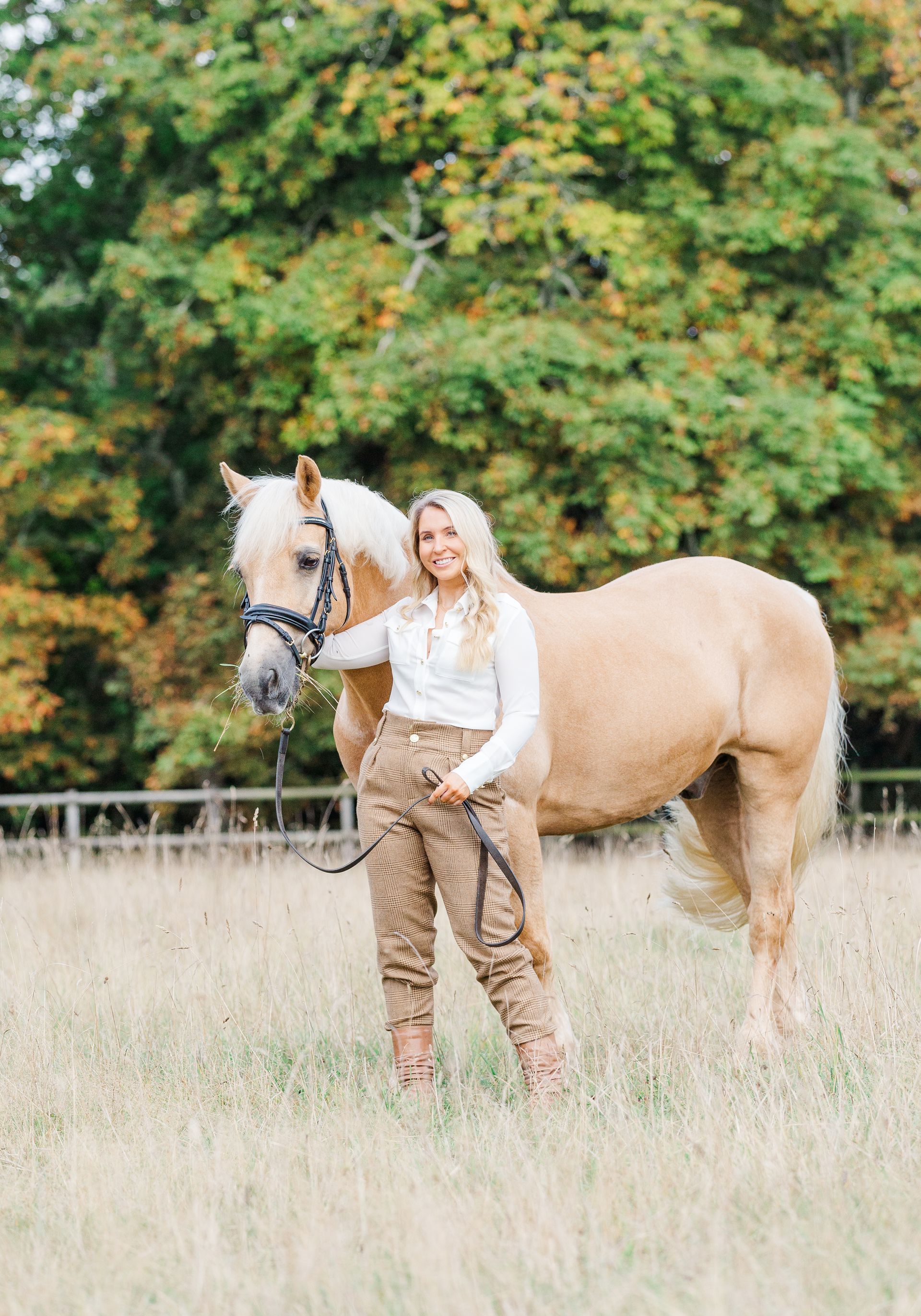 Woman in riding attire poses with a palomino horse in a grassy field.
