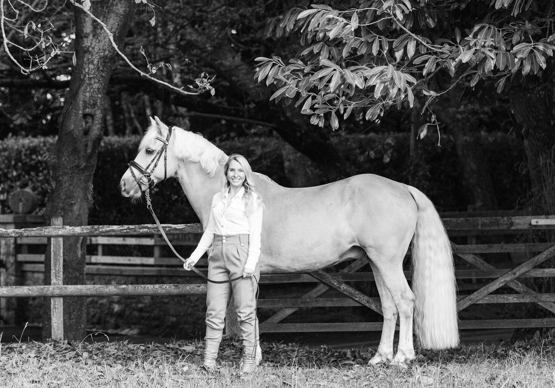 Woman standing beside a palomino pony in a field, holding its lead rope near a wooden fence and trees.