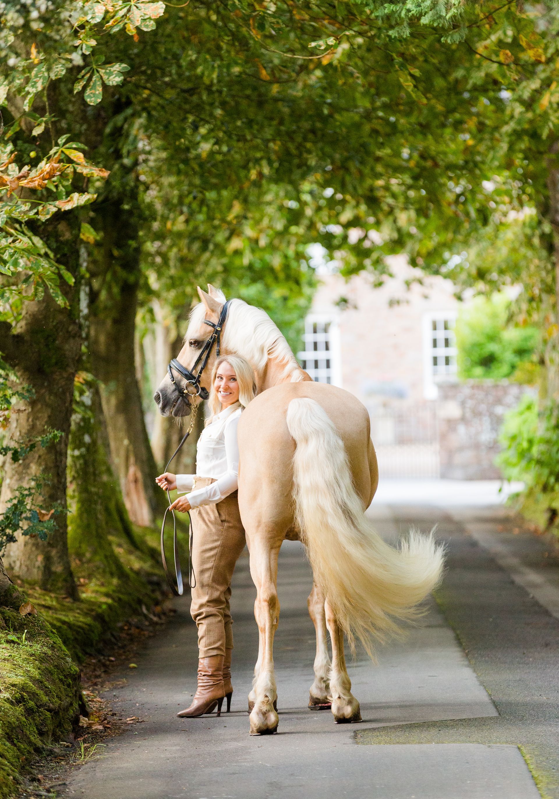 Woman in tan pants and white top stands with a palomino horse on a paved path, trees overhead.