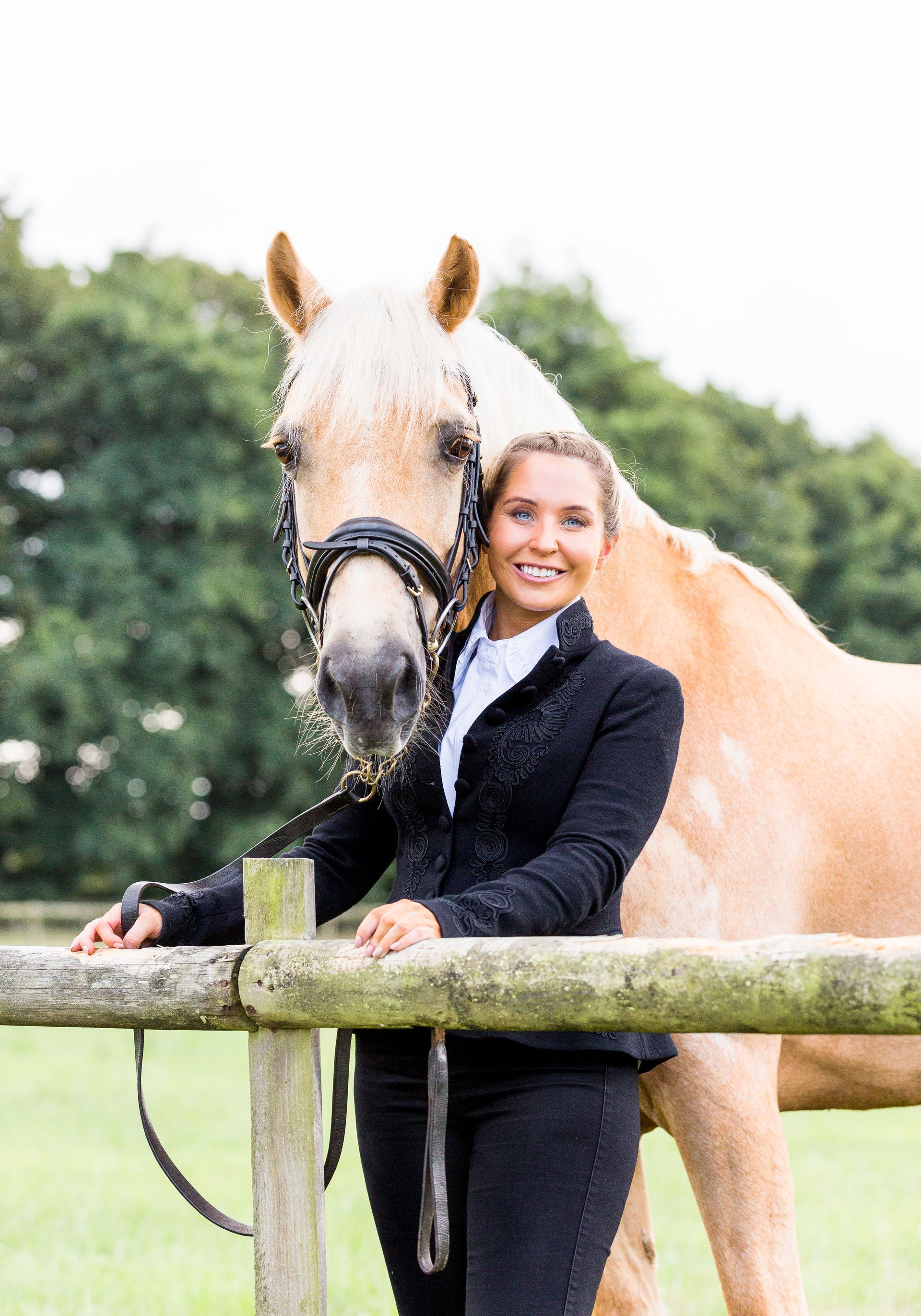 Woman in riding attire with a tan horse, leaning on a fence, smiling outdoors.