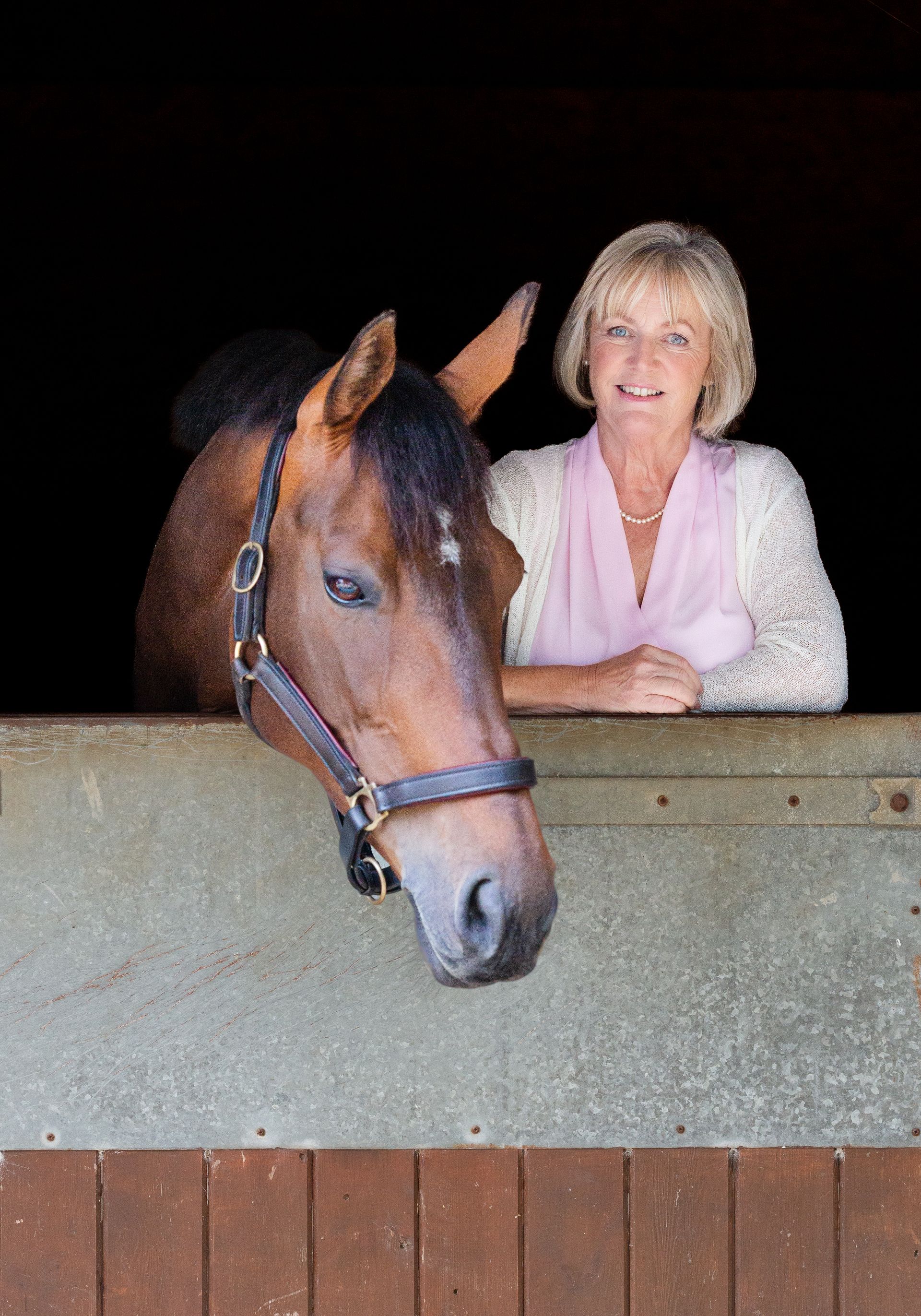 Woman and brown horse in a stable. Woman smiles, rests arm on stall. Horse looks toward the viewer.