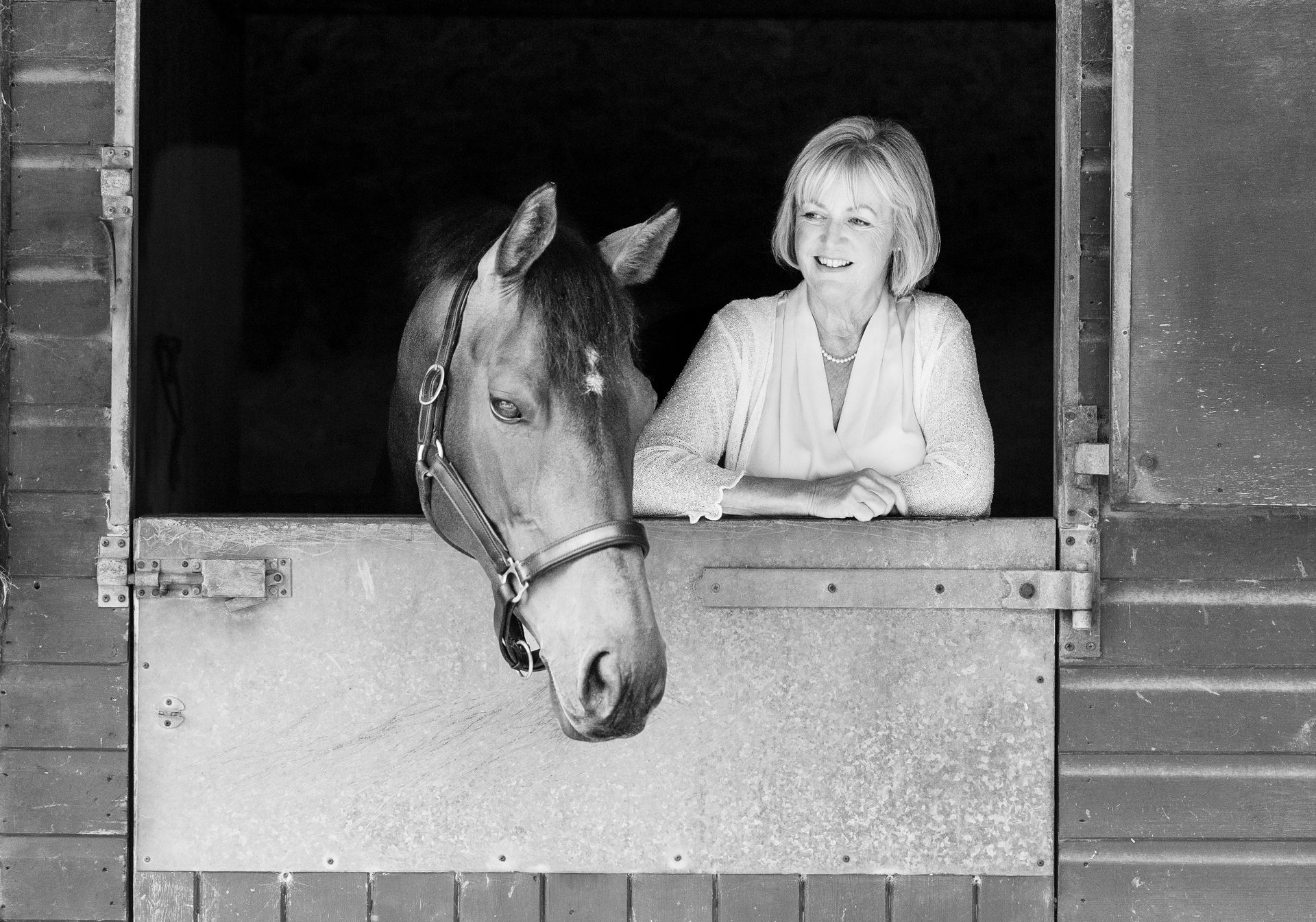 Woman and horse in a stable doorway. The woman smiles, resting arms on the door. The horse peers out.