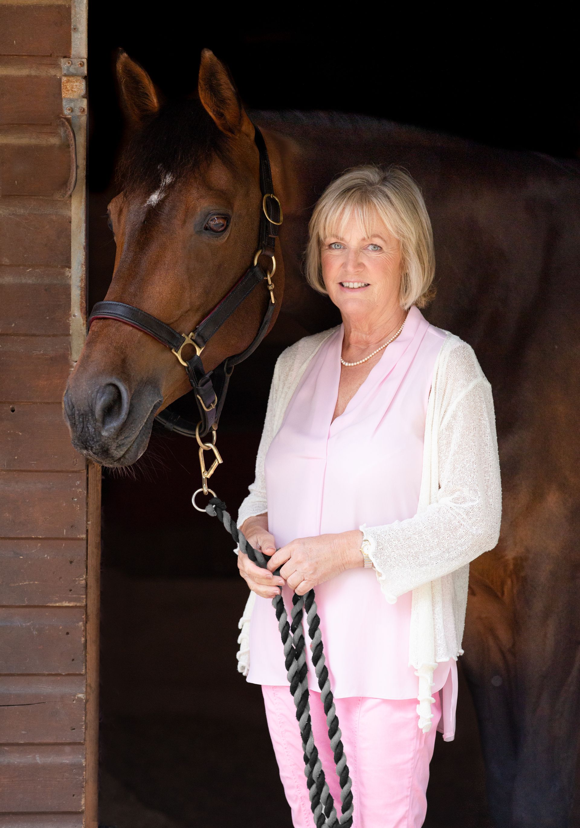 Woman in pink and cream with a brown horse in a stable doorway.