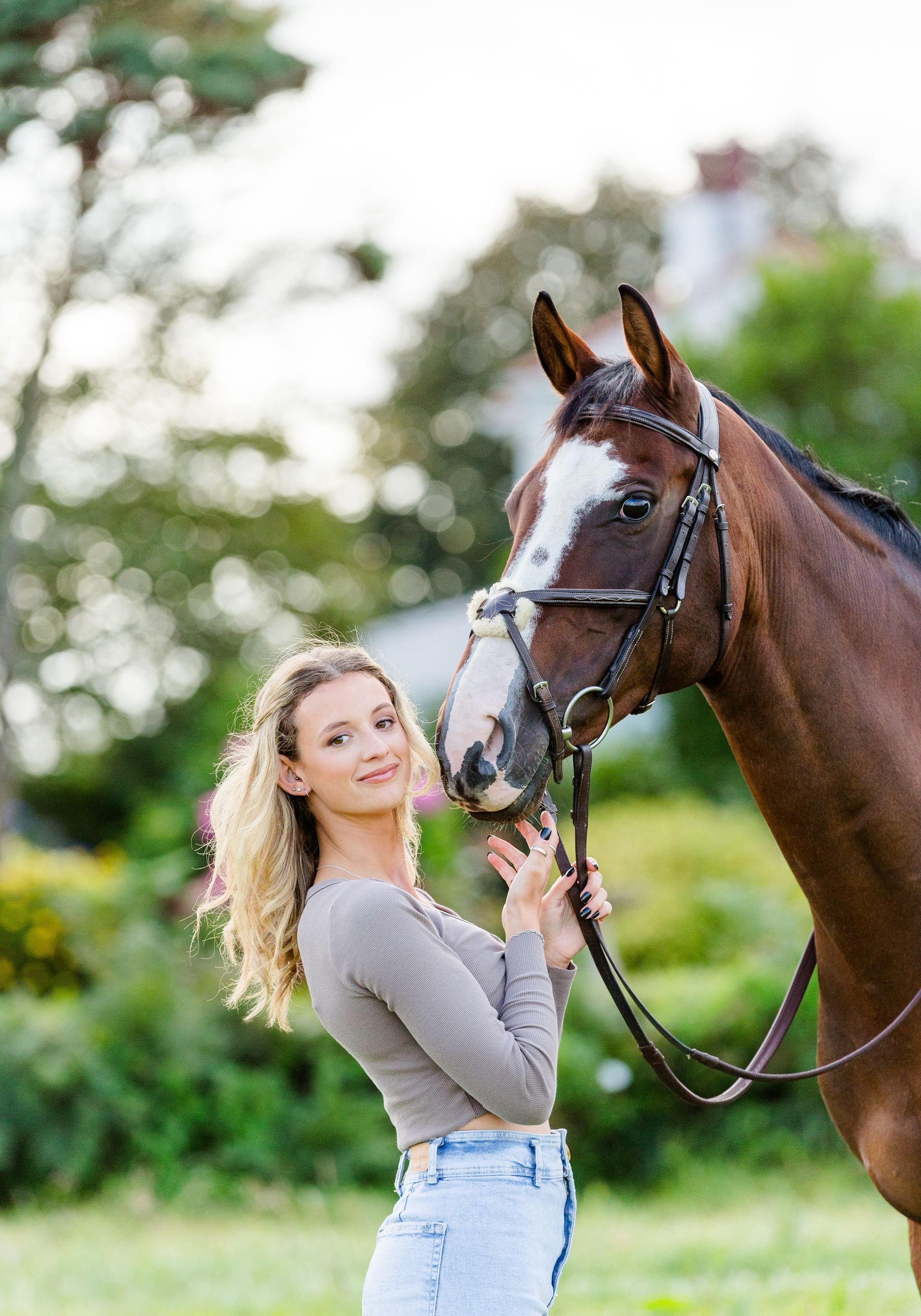 Woman in crop top and jeans smiles at a brown horse with a white face in a grassy field.