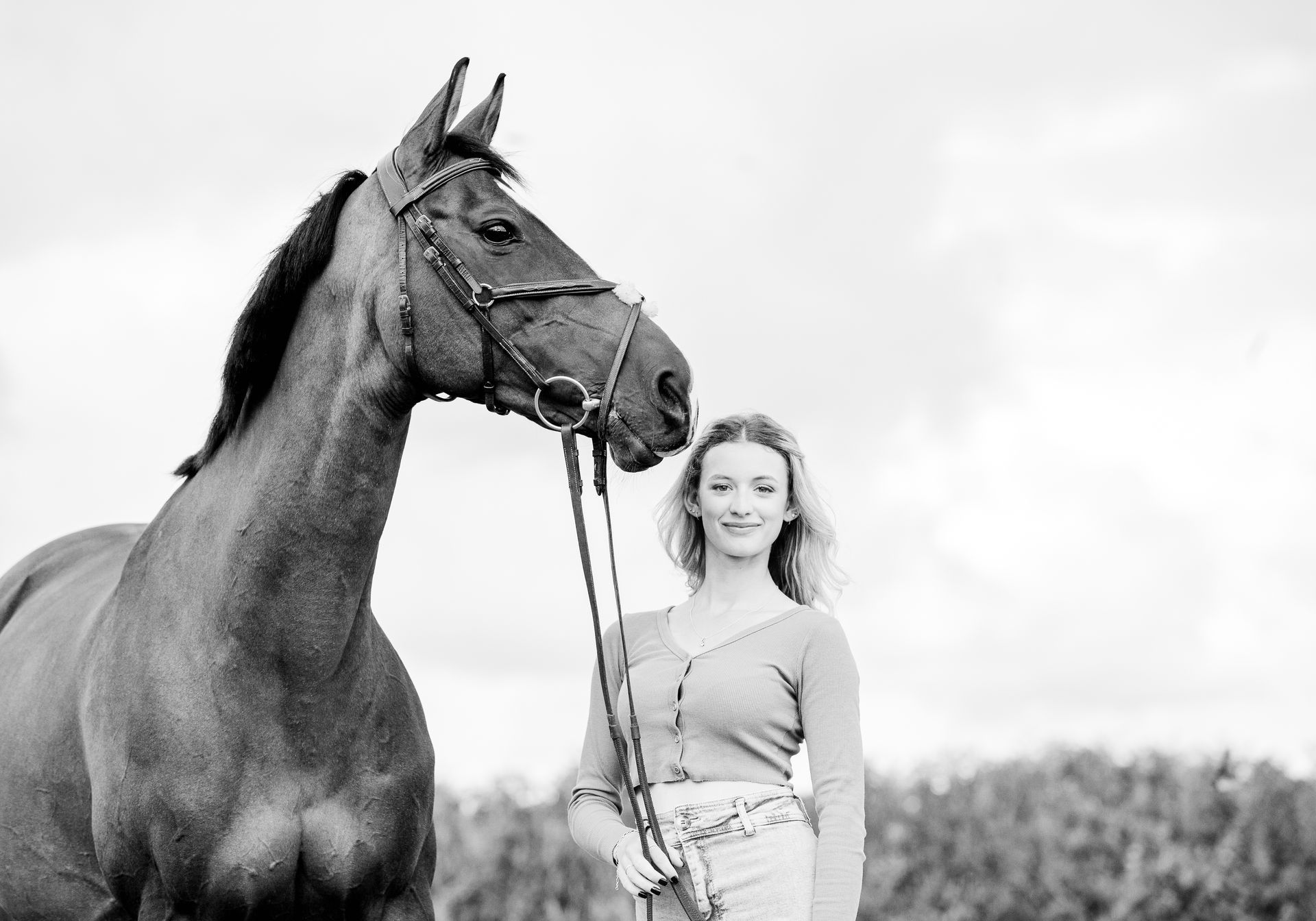 Woman in cardigan stands with a horse, holding its lead rope. Cloudy sky in the background.