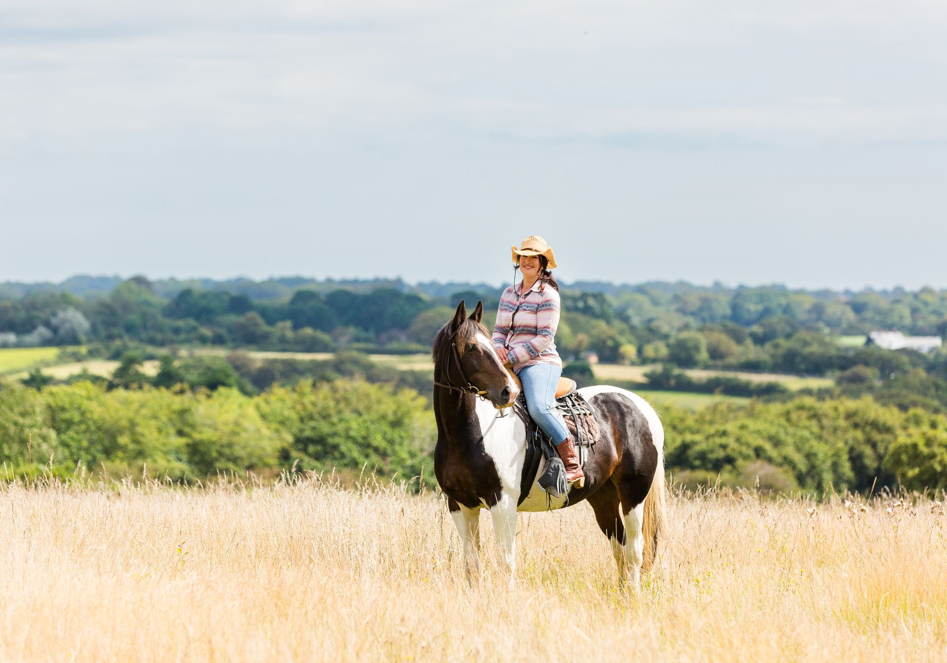 Woman in cowboy hat on a painted horse in a grassy field overlooking a green landscape.