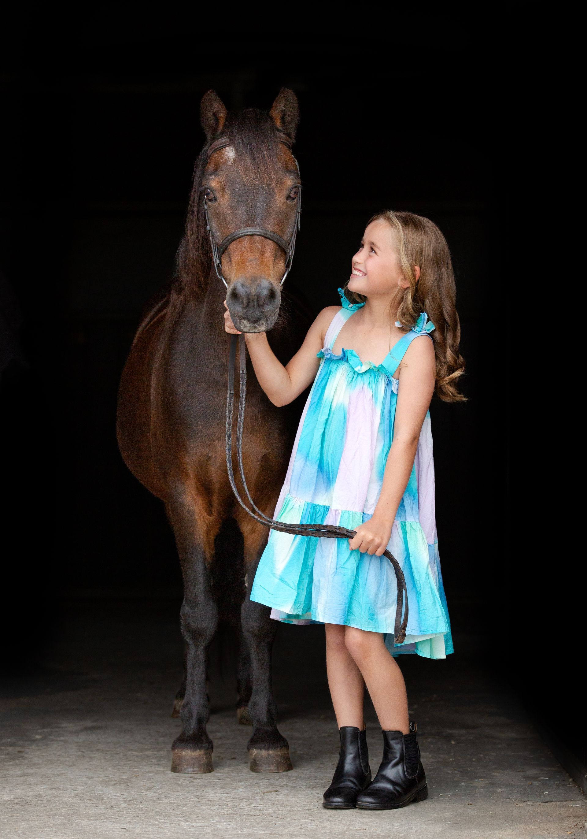 Young girl in a blue dress smiles, holding a horse's reins in a dark stable.