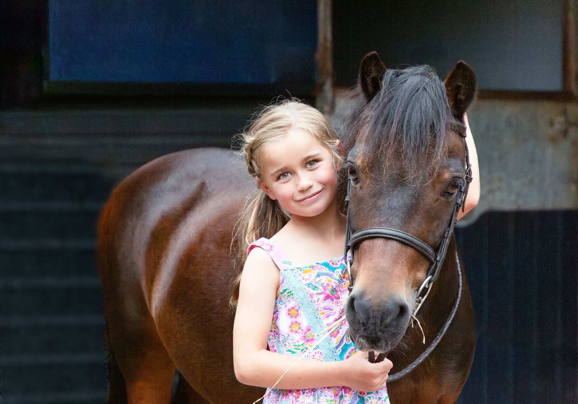 Girl smiles, hugging a brown pony with a dark mane, in front of a shadowed building.