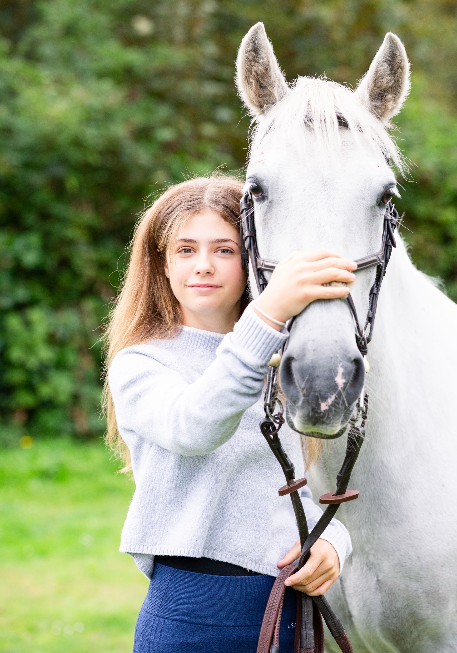 Girl in blue sweater and jeans with white horse, outdoors, holding horse's bridle.