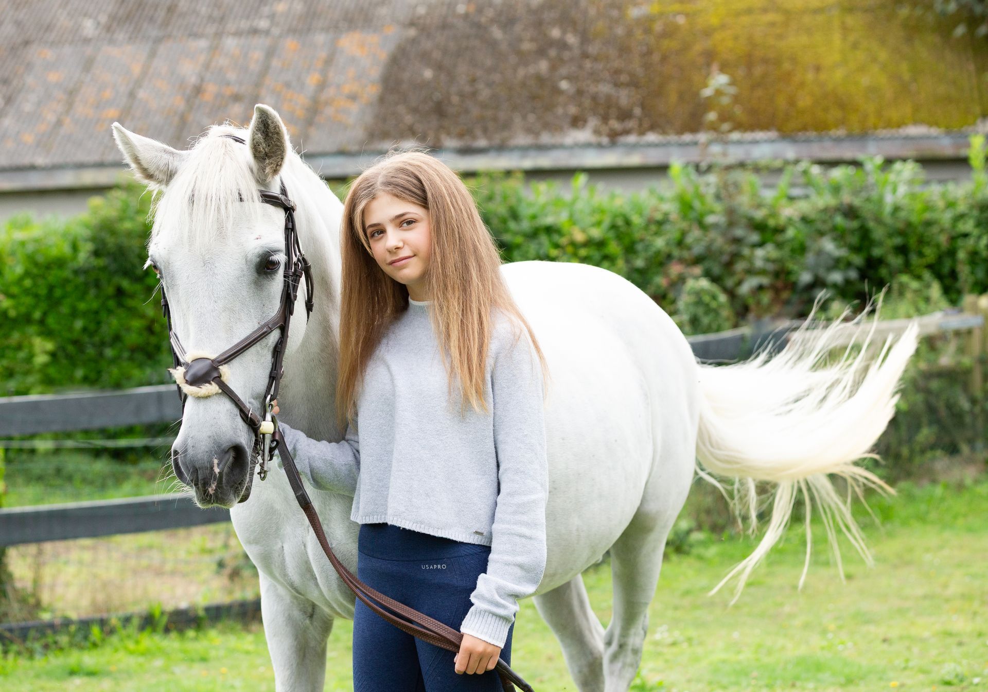 Young person with long curly hair stands next to a white horse, both in a grassy outdoor setting.